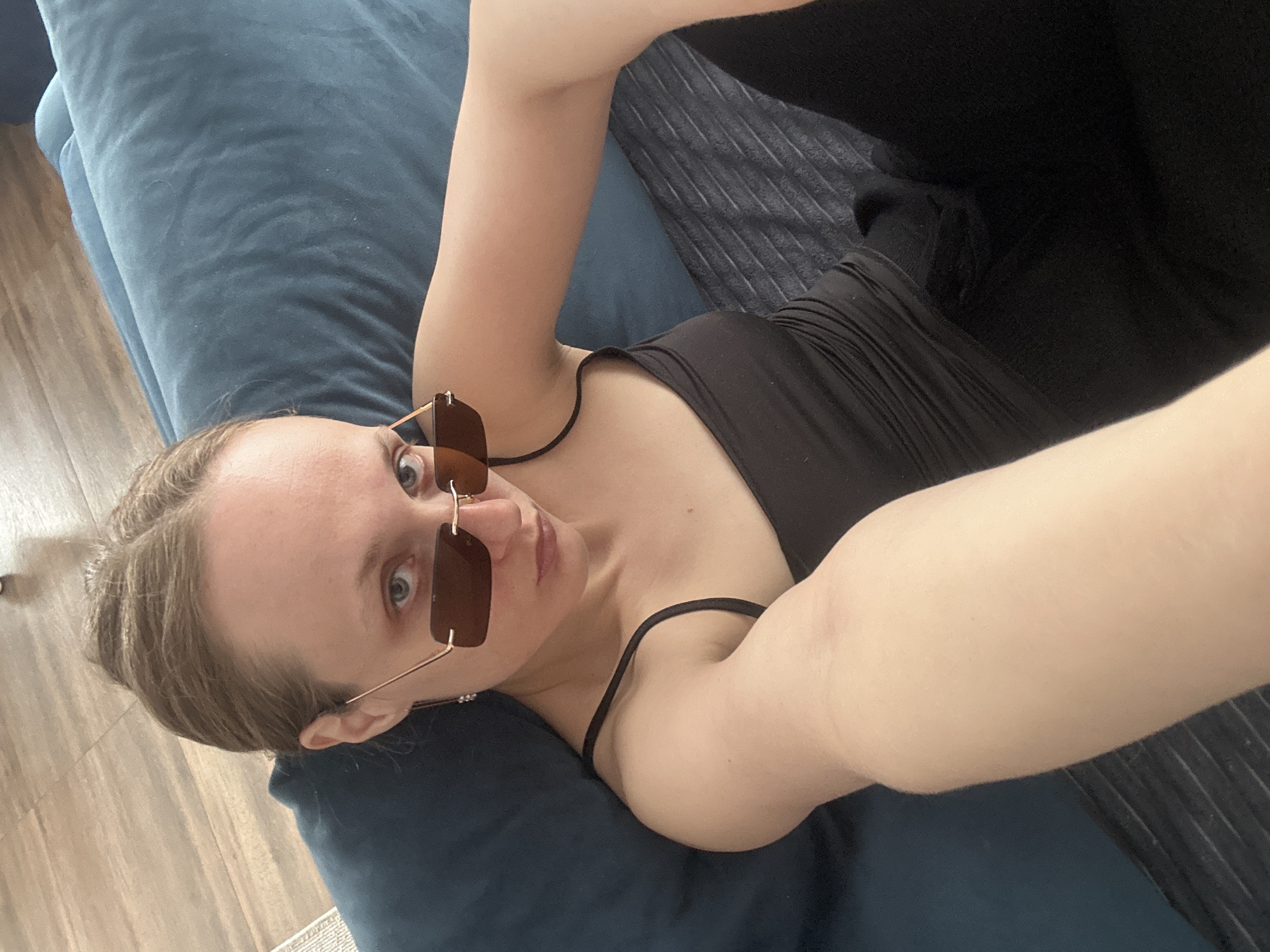 A selfie shows a fair-skinned woman with her hair pulled back, wearing brown-tinted sunglasses and a black top, lying on a blue velvet couch. She is looking directly at the camera. The couch is placed on a wooden floor, and a blanket is visible to the right of her body. There is not enough information to assess the location in the city of Sepych, Russia.
