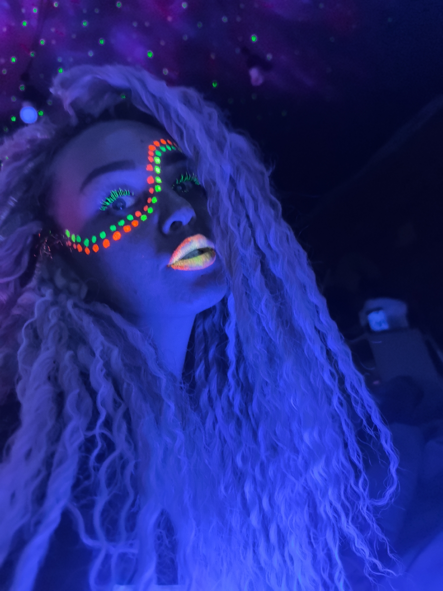 A person is depicted in a close-up portrait, illuminated by blacklight (UV light), creating a vibrant and glowing effect. The overall scene is dark, cast in deep blue and purple tones from the UV lighting.
The subject's face is adorned with elaborate fluorescent makeup. Small, circular dots of glowing orange and green are intricately patterned across her forehead and cheekbone, extending towards her right eye. Her eyelashes are a prominent feature, glowing intensely in green and appearing long and wispy. Her lips are also luminescent, showcasing a vibrant gradient of yellow and orange. The UV light gives her skin a subtle bluish-purple tint.
Her hair, which appears long, wavy, and voluminous, glows with a strong blue-purple hue under the UV light, cascading down the left side of the frame. The subject is looking upwards and slightly to the right, with a neutral to contemplative expression.
The background is dark and out of focus, featuring blurred green dots and hints of purple light, suggesting an indoor environment like a club, party, or performance setting designed with UV effects. No specific location cues beyond the general ambiance are visible to pinpoint a precise spot in Noviy, Russia. There is no visible text in the image.