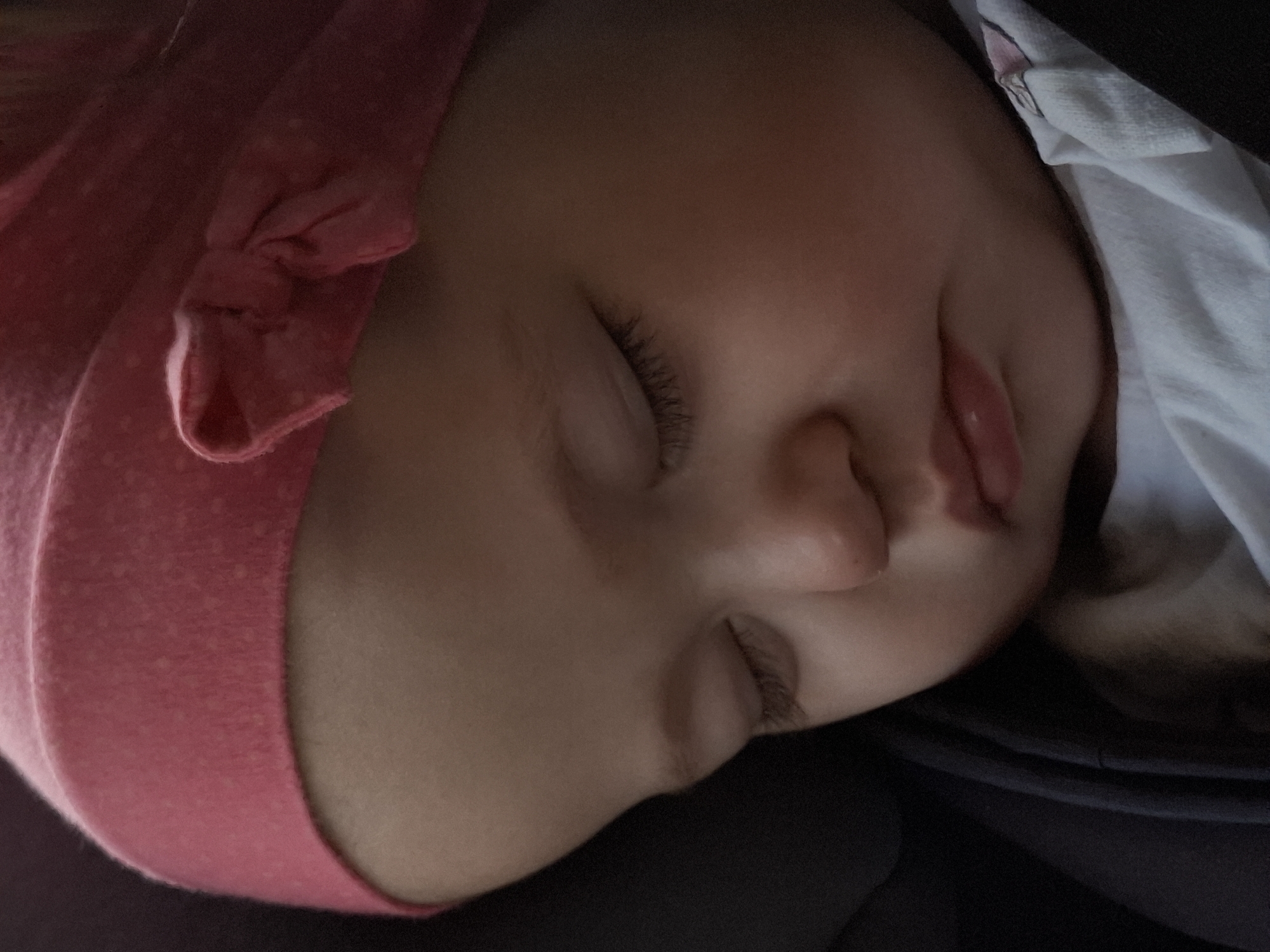 A close-up image shows a sleeping baby. The baby is wearing a pink hat with a bow. The baby's face is tilted, and their eyes are closed. A white cloth is draped near the baby's face. The background is dark. There is no information about the location in the city of Chaikovsky, Russia.