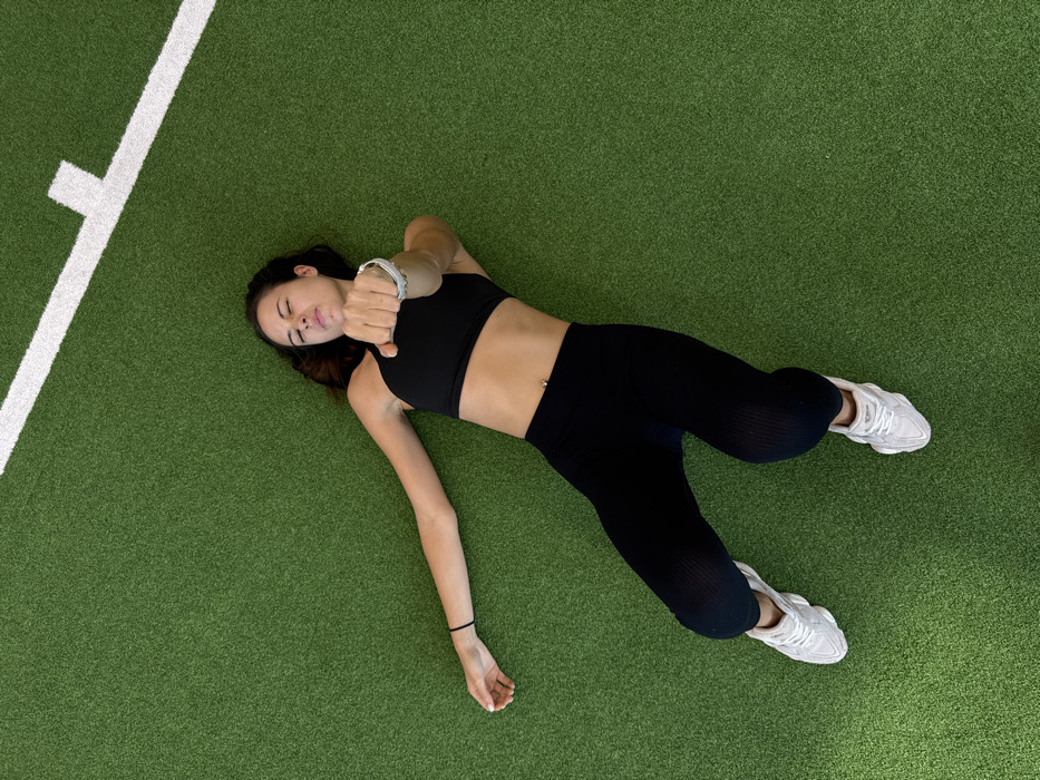 A single female is depicted lying supine on a green artificial turf surface, viewed from an overhead perspective. She wears a black sports bra, black ribbed capri-length leggings, and white athletic shoes. A silver navel piercing is visible. Her left arm is extended straight, while her right arm is bent at the elbow, with the hand clenched into a fist and extended towards the camera. Her legs are bent at the knees, with feet positioned flat on the turf. Her facial muscles are contracted, with closed eyes and a slightly open mouth. A white line, forming a corner, is visible on the green turf in the upper right portion of the frame. The location is Bangkok, Thailand.