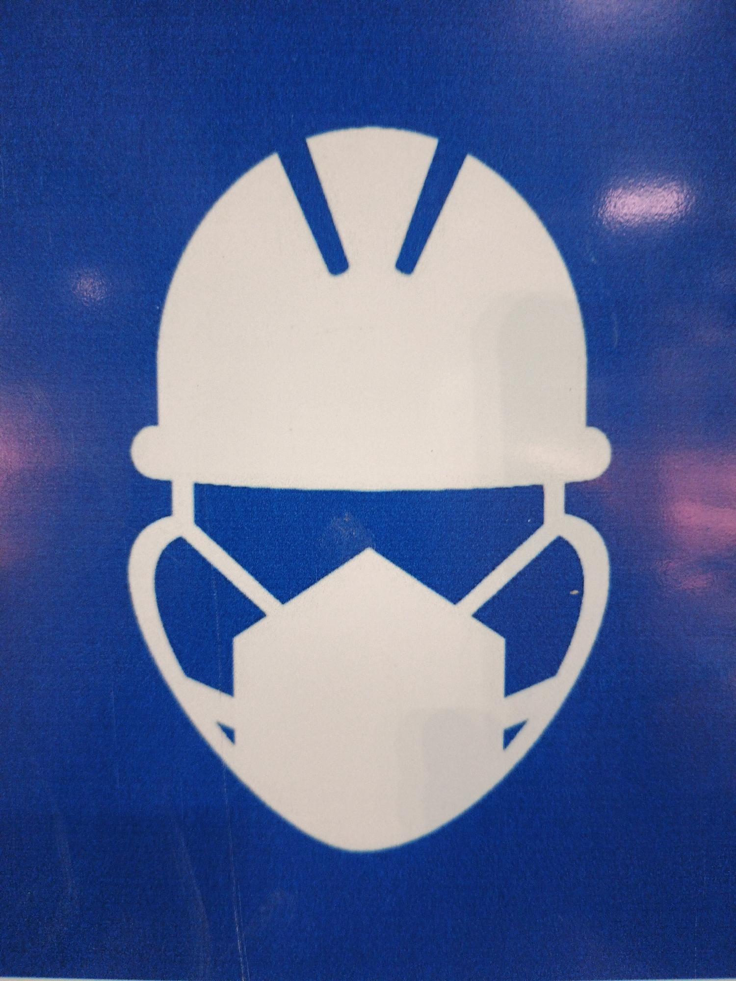 The image is a close-up shot of a safety sign with a white pictogram on a blue background. The pictogram depicts a stylized head wearing both a hard hat and a face mask. The hard hat is white and has a rounded top with two angled lines representing ventilation or design features. Below the hard hat, a white mask covers the lower half of the face, extending to the sides with straps that loop around the ears. The mask has a slightly angled, geometric shape over the nose and mouth area. The blue background of the sign has some subtle texture and faint, possibly reflective, pinkish light streaks, suggesting an indoor environment with artificial lighting. The location of this sign within Lolawang, Indonesia, is not discernible from the image itself.