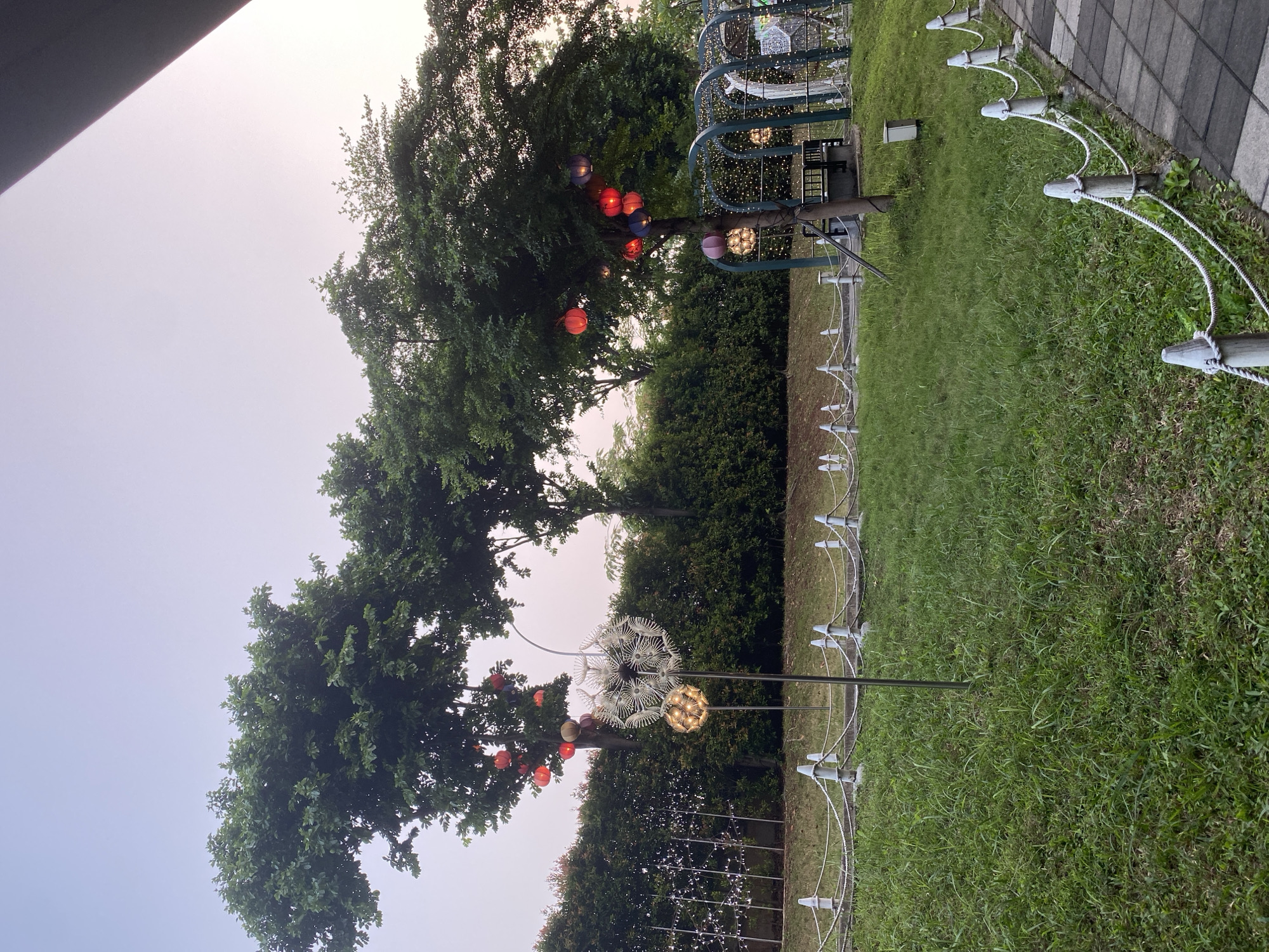 This media file depicts an outdoor scene at dusk, likely in a park or recreational area, given the presence of lush green grass, trees, and decorative lighting. The sky is overcast, suggesting a cloudy or hazy atmosphere, typical of the twilight hours. The image is oriented vertically, with the ground and grassy area occupying the right side and the sky and tree canopies on the left.
The primary subjects are the natural elements and decorative objects. Several trees with dense green foliage are visible, some adorned with colorful lanterns. These lanterns, in shades of red, blue, and purple, hang from the branches, creating a festive ambiance. In the background, a large, arched structure is illuminated with numerous small, white lights, resembling a string of fairy lights. There are also two prominent decorative lights resembling dandelions on a pole. One is a large, white, intricate sphere, and the other is a smaller, golden sphere with visible lights.
In the foreground, a pathway made of checkered grey and black tiles runs along the edge of the grassy area. A decorative rope fence, supported by short wooden posts with white caps, separates the pathway from the grass. The fence appears to be made of white rope tied to the posts.
The scene suggests a public space that is being decorated for an event or to enhance its nighttime appeal. The time of day is clearly evening, as indicated by the dim sky and the illuminated decorations. The overall mood is calm and slightly magical due to the lanterns and string lights. There is no visible text in the image. The location context provided, East Jakarta, Indonesia, suggests a tropical or subtropical climate where such outdoor decorations and lush greenery are common. No people are visible in the frame.