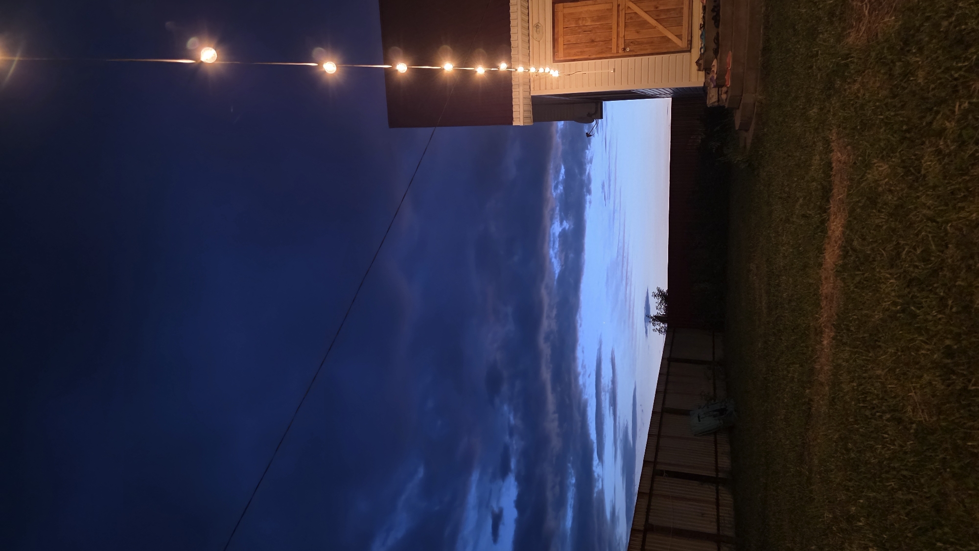 The image shows a backyard at dusk. A chain of lights hangs from a wire strung across the frame, illuminating a section of a light-colored house with a dark roof and a wooden door. The house appears to be a shed or outbuilding. A wooden fence runs along the left side of the yard, with a patch of grass and some shadows in the foreground. The sky is a deep blue with dark, cloudy streaks, suggesting an approaching storm or the end of daylight. The overall impression is one of a quiet, rural or suburban setting.