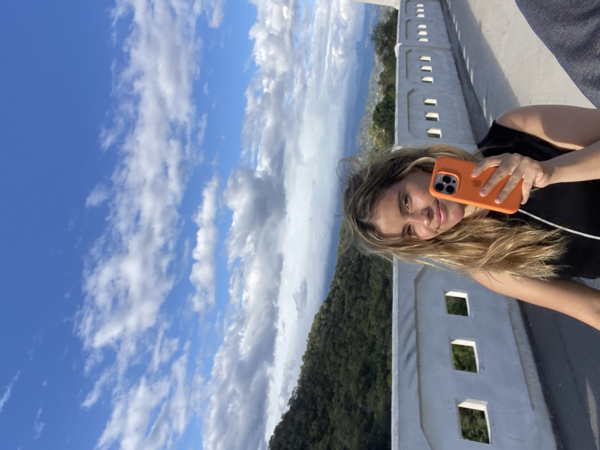 A woman is holding an orange cell phone in her right hand, taking a selfie on a bridge. The backdrop features a landscape including a hillside covered in trees, mountains in the distance, and a sky filled with clouds. She is standing near a white concrete barrier with square openings. The location is San Fernando del Valle de Catamarca, Argentina.
