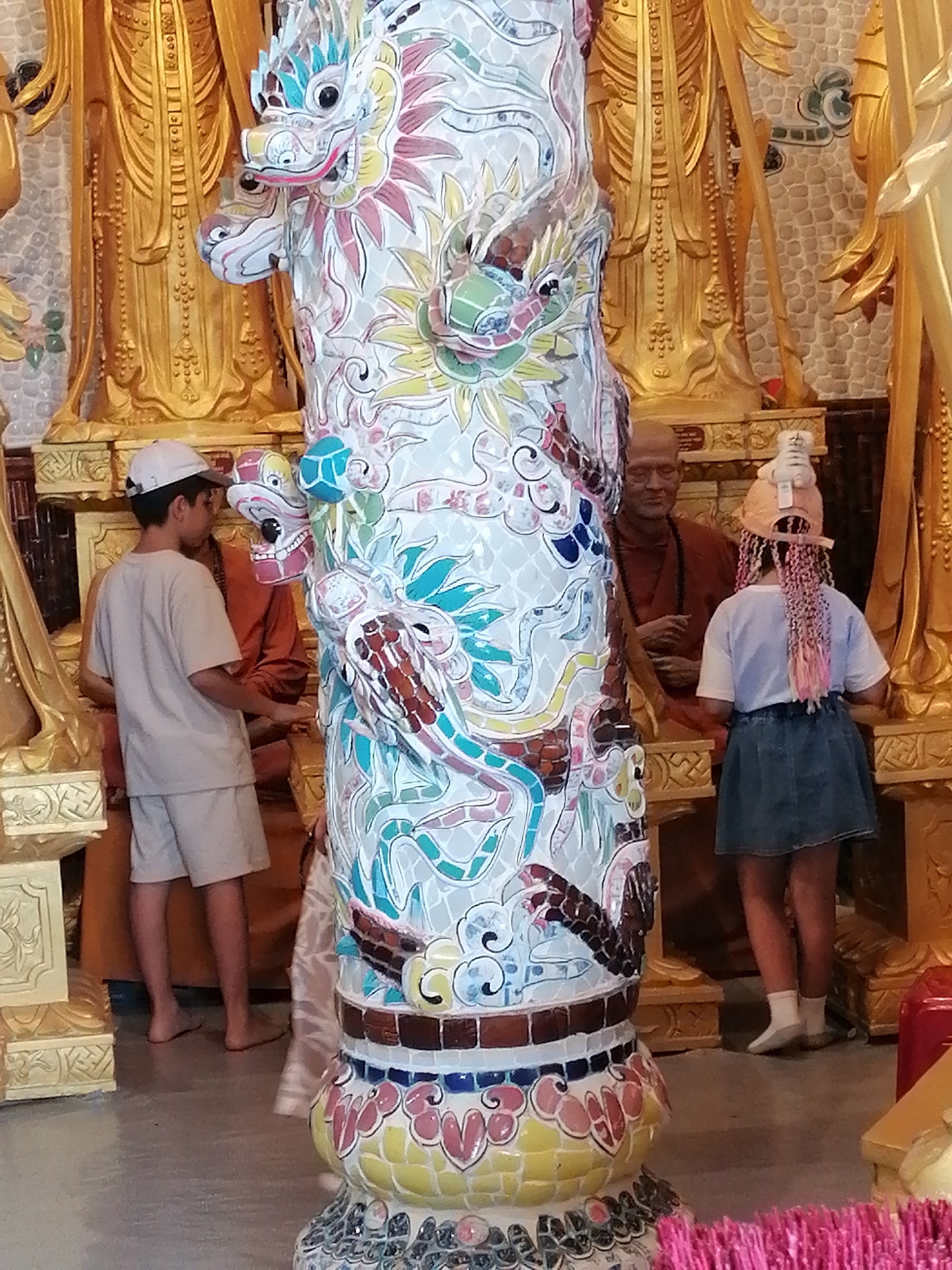This is a full-body, eye-level, medium shot of a colorful ceramic dragon statue in front of golden Buddhist statues in a temple. In the foreground, the dragon statue is the central focus, covered in mosaic tiles depicting dragons and floral patterns. To the left of the statue, a young boy wearing a white cap, a light-colored t-shirt, and shorts stands facing away from the camera. Behind the boy, a person in an orange robe is partially visible. To the right of the statue, a young girl with pink braids, a white t-shirt, and a denim skirt stands facing the dragon statue. She has a small stuffed animal attached to her hat. The background consists of large, ornate golden statues of Buddhist deities, characteristic of temples in Vietnam, specifically in Lộc Quý, Vietnam, where this scene is likely situated. The floor appears to be polished concrete.