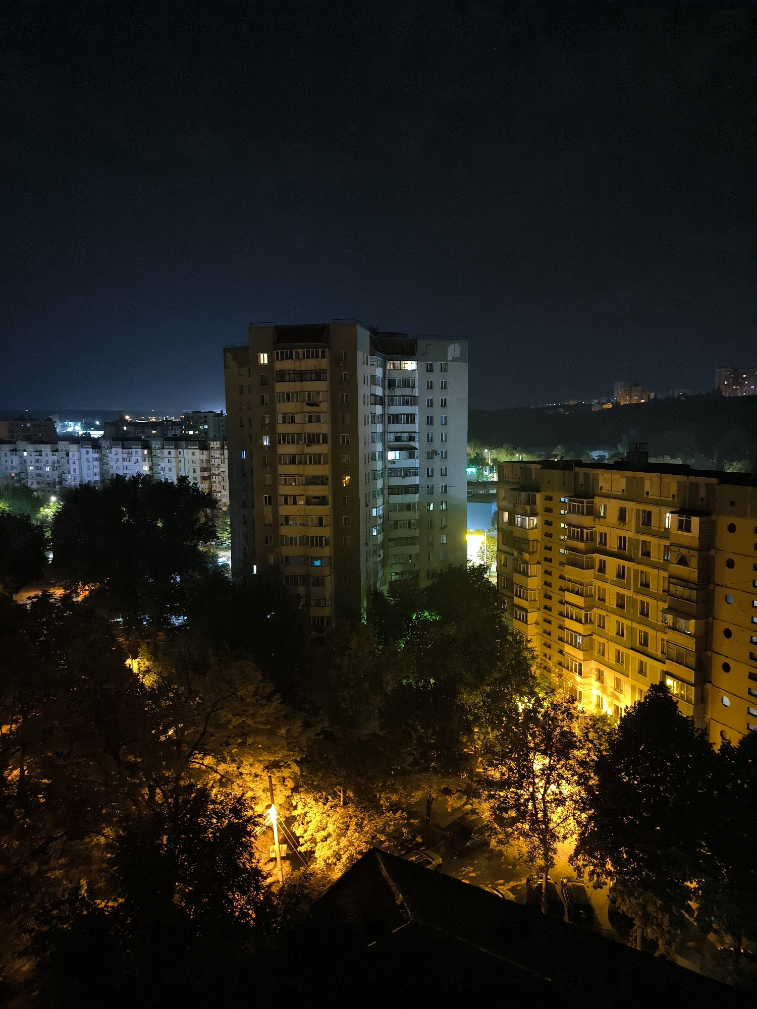 The image is a nighttime aerial view of a city, likely in Chișinău, Moldova, characterized by apartment buildings and streetlights illuminating trees. In the foreground, the dark silhouettes of trees are prominent, with bright orange streetlights casting an intense glow through their leaves. Beyond the trees, several multi-story apartment buildings are visible. The tallest building, on the right, is a yellow-toned structure with numerous windows, many of which are lit. To its left, a slightly taller, gray apartment building also shows illuminated windows. Further in the background, a series of similar apartment buildings stretch across the landscape, with various windows lit in different colors, indicating habitation. The night sky is dark blue, and in the distance, the faint outlines of more buildings and a hilly terrain can be seen. The overall impression is of a residential urban area at night, with artificial lighting dominating the scene.