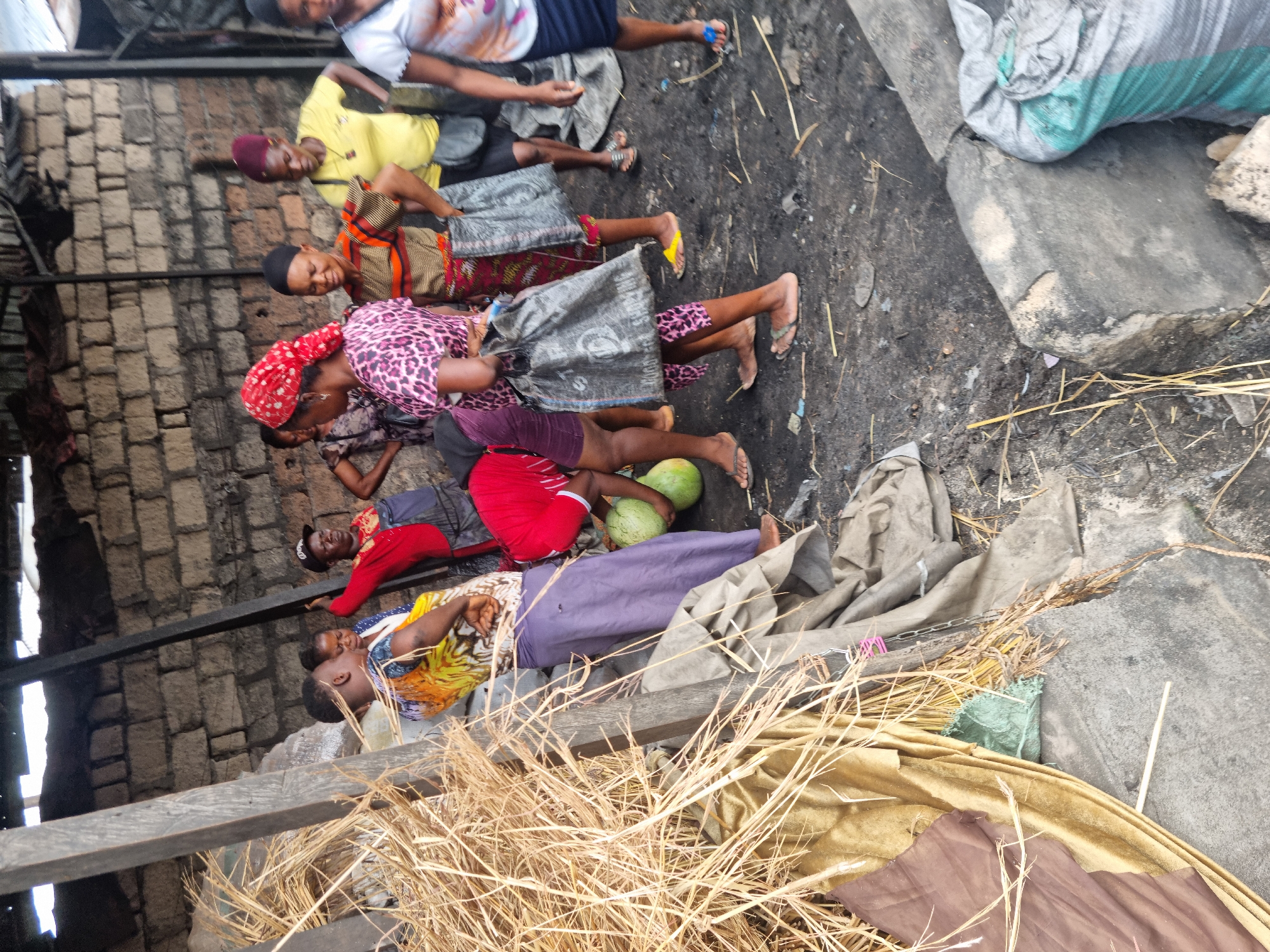 The image captures a group of approximately ten individuals, predominantly women and younger adults, resting or sitting on the ground in an outdoor setting. The scene is shot from a slightly elevated perspective, looking down at the people.
The subjects are arranged in various relaxed postures, mostly sitting or lying on the dark, uneven ground which is scattered with dry straw, small debris, and what appears to be charcoal or ash. Several individuals wear casual clothing, including patterned dresses, skirts, shorts, and t-shirts, with some women having head coverings. Their footwear varies, including sandals and bare feet.
In the mid-ground, two green watermelons are visible near a person lying down, suggesting the context might involve selling produce or taking a break from carrying goods. Fabric bags, some appearing to be made of coarse material, are also scattered around, some seemingly used as makeshift seats or pillows.
The background on the left features a rough brick wall, indicating a building or structure. Wooden beams run across the foreground and mid-ground, possibly part of a simple shelter or a market stall. The lighting suggests it is daytime. No discernible text is visible on any objects or surfaces. The overall atmosphere appears to be one of rest or waiting in a busy, informal environment.