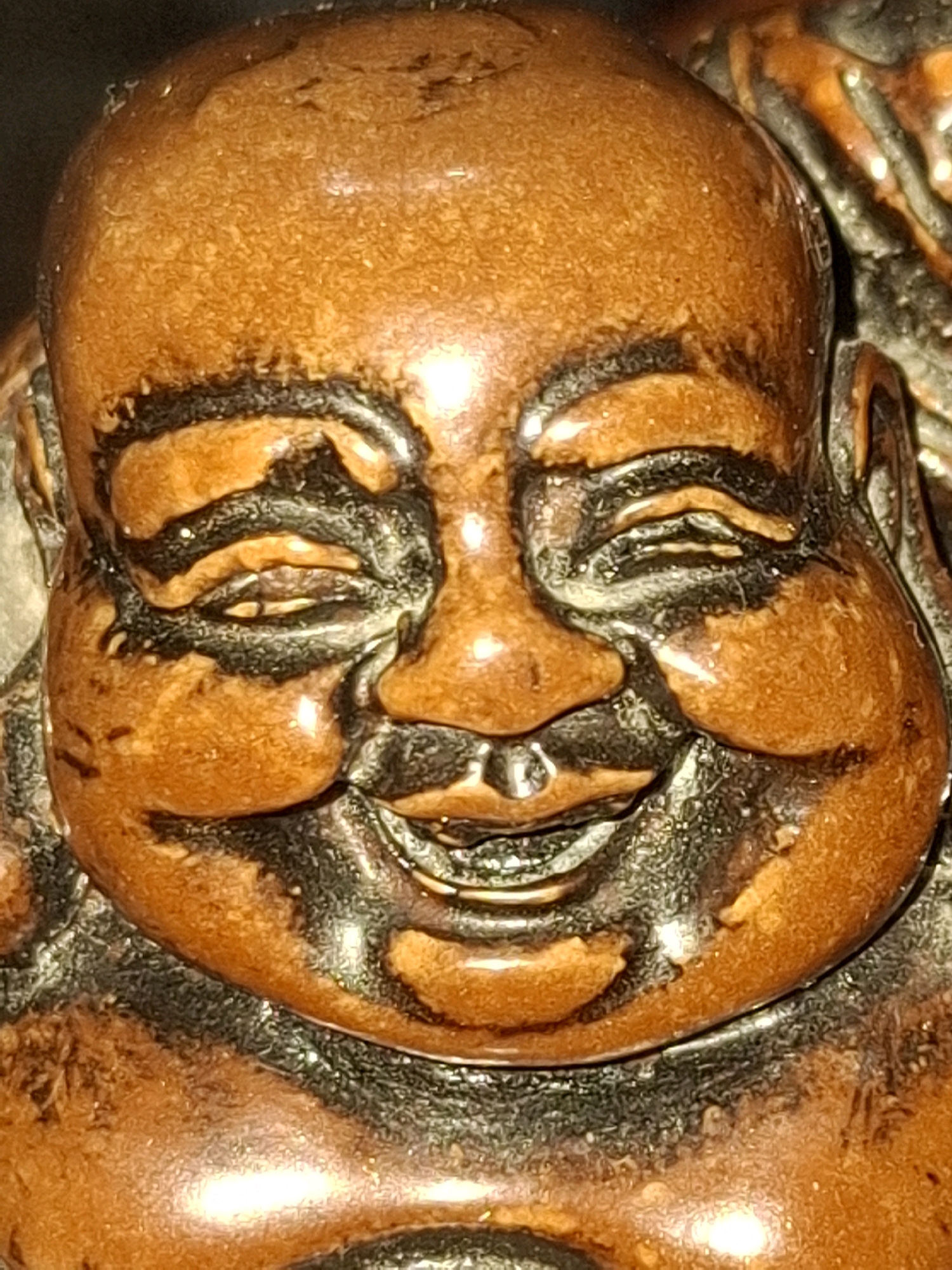 The image is a close-up, frontal view of a carved wooden figurine of a laughing Buddha. The figurine is predominantly a reddish-brown color with darker, possibly black, carved details around the eyes, nose, and mouth. The surface of the wood appears smooth and slightly glossy, with some visible texture. The figurine's face is round and features prominent, rounded cheeks, a wide smile revealing teeth, and crinkled eyes, all contributing to an expression of joy. The lighting creates highlights on the surface of the wood, particularly on the forehead and cheeks. The background is dark and out of focus. This image does not depict any identifiable location in Almaty, Kazakhstan, nor does it contain any people or indicate any specific actions or interactions. It is a still, focused shot of an inanimate object.