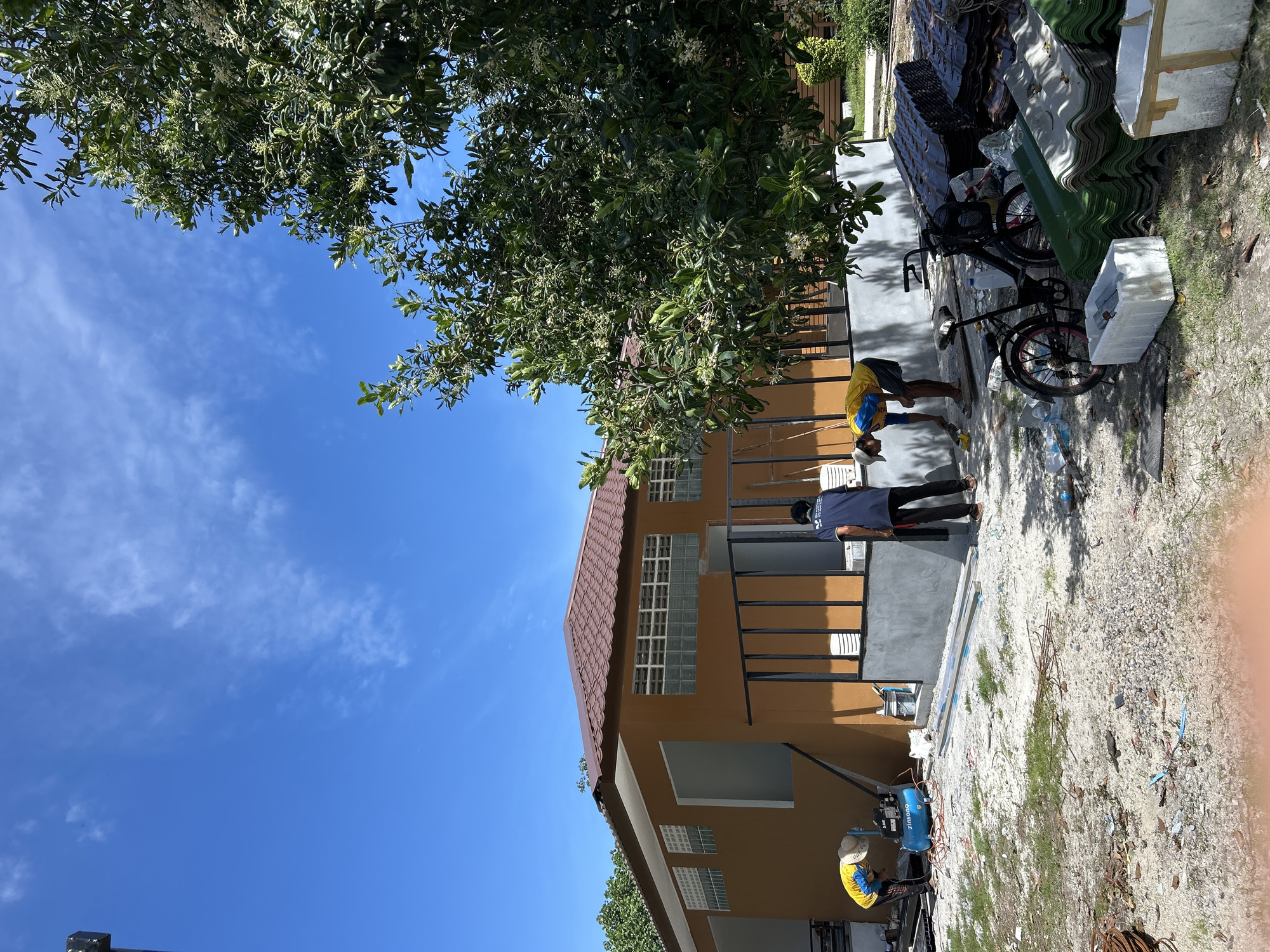 The image captures an outdoor construction or renovation site in Ao Nang, Thailand, under a clear blue sky with scattered white cirrus clouds. A single-story building, painted an orange-brown hue with a brown tiled gabled roof, is the primary structure. Its upper section features horizontal rows of glass block windows and open rectangular voids, while the lower front wall exhibits a freshly applied light grey concrete finish and a dark metal railing.
Three male workers are present. On the left, a worker in a yellow and blue shirt, dark shorts, and a light-colored hat is bent over, attending to a blue "zinsano" air compressor resting on the gravel ground. A second worker, wearing a dark blue shirt and dark pants, stands near the grey wall section, facing away from the camera and holding a dark, slender object. A third worker, dressed in a yellow and blue shirt, is bent over, actively working on the grey lower wall surface.
The ground consists primarily of light-colored gravel and dirt, interspersed with sparse green grass and various construction debris, including coiled wires, small pieces of material, and a white plastic bucket. To the right, numerous stacks of corrugated roofing sheets are visible, with dark brown sheets piled atop green ones. Two white styrofoam boxes are also present near these materials. A black bicycle with pink accents on its wheels and seat, and a front basket, is parked among the stacked materials. A large, leafy green tree with small white blossoms dominates the right side of the frame, partially obscuring the building and casting shadows. Further green foliage is visible in the background.