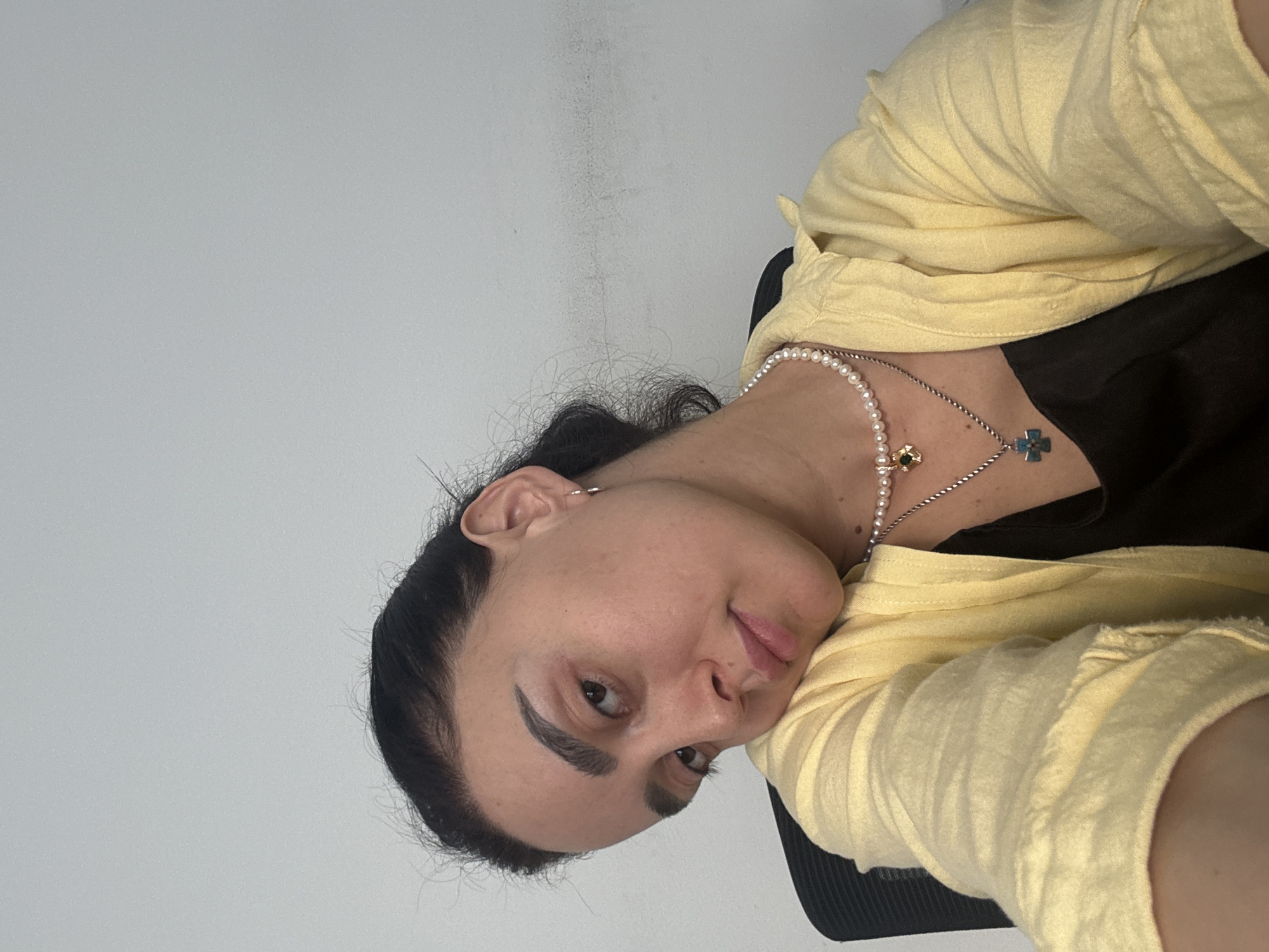 A woman is seated indoors, possibly in an office. She is wearing a yellow shirt over a black top, and a necklace. The background is a plain white wall with some minor stains. The image was likely taken in Tbilisi, Georgia.