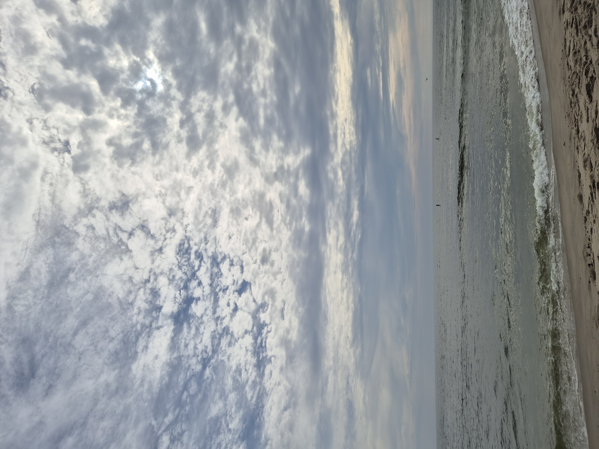 An overcast day at the beach in Middletown Township, USA, with a wide expanse of ocean meeting a sandy shore. The sky is filled with a textured pattern of gray and white clouds, with patches of sunlight breaking through in the upper right quadrant. The ocean is a muted green-gray, with gentle waves lapping at the shoreline, creating a foamy white edge. In the distance, a few indistinct figures can be seen on paddleboards in the water, and a bird is flying in the sky. The sand in the foreground is light brown, marked with footprints and small pebbles. The scene evokes a sense of calm and natural beauty.