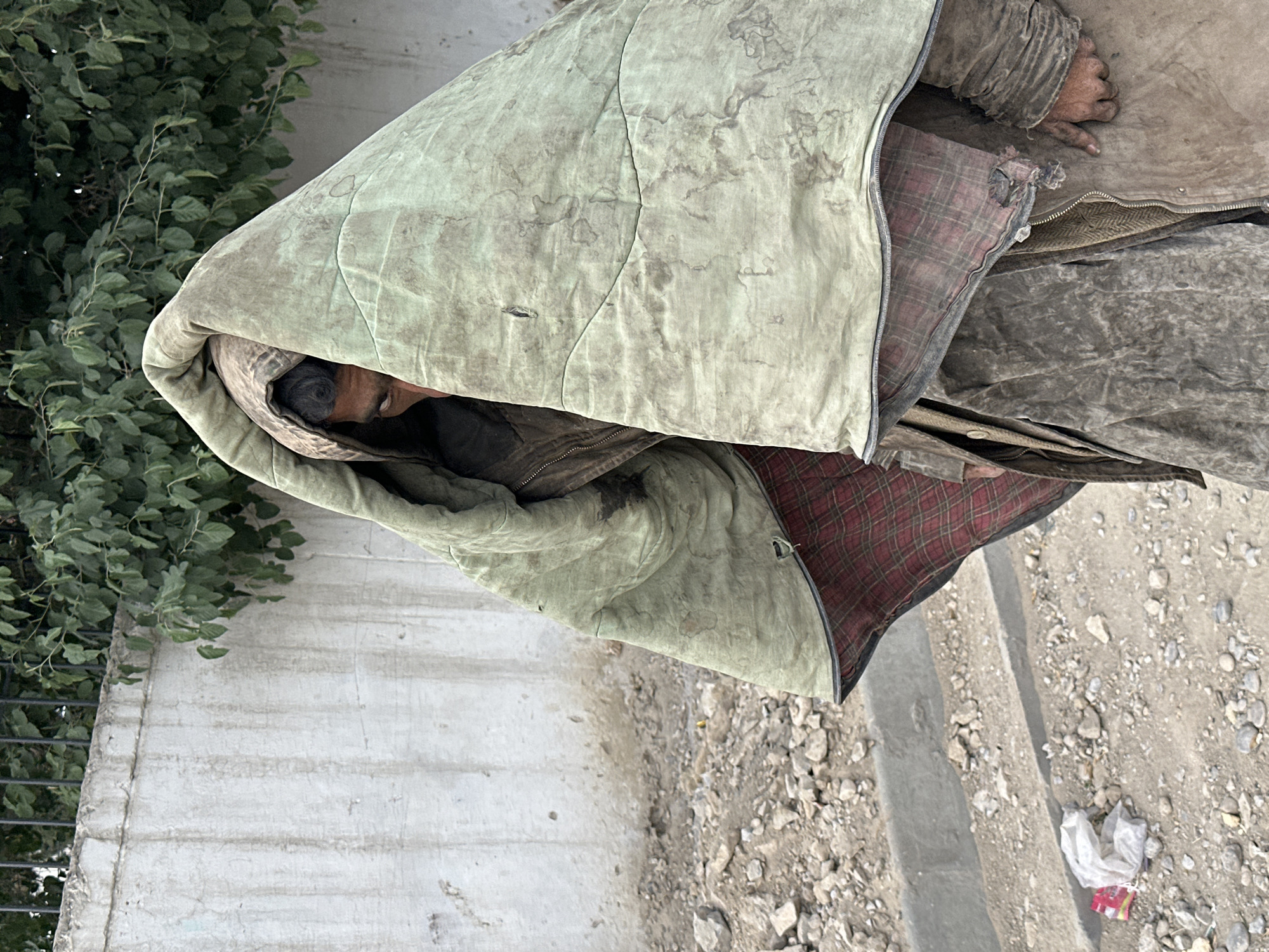 A homeless individual is seen on a street in Quetta City Tehsil, Pakistan. The person is wrapped in a dirty, light-green quilted blanket. The individual is lying on the ground next to a concrete wall and a green leafy plant. Patches of dirt, rocks, and trash surround the location. The person is lying down, partially covered by a plaid fabric. Their face is visible, and their hand is slightly exposed.
