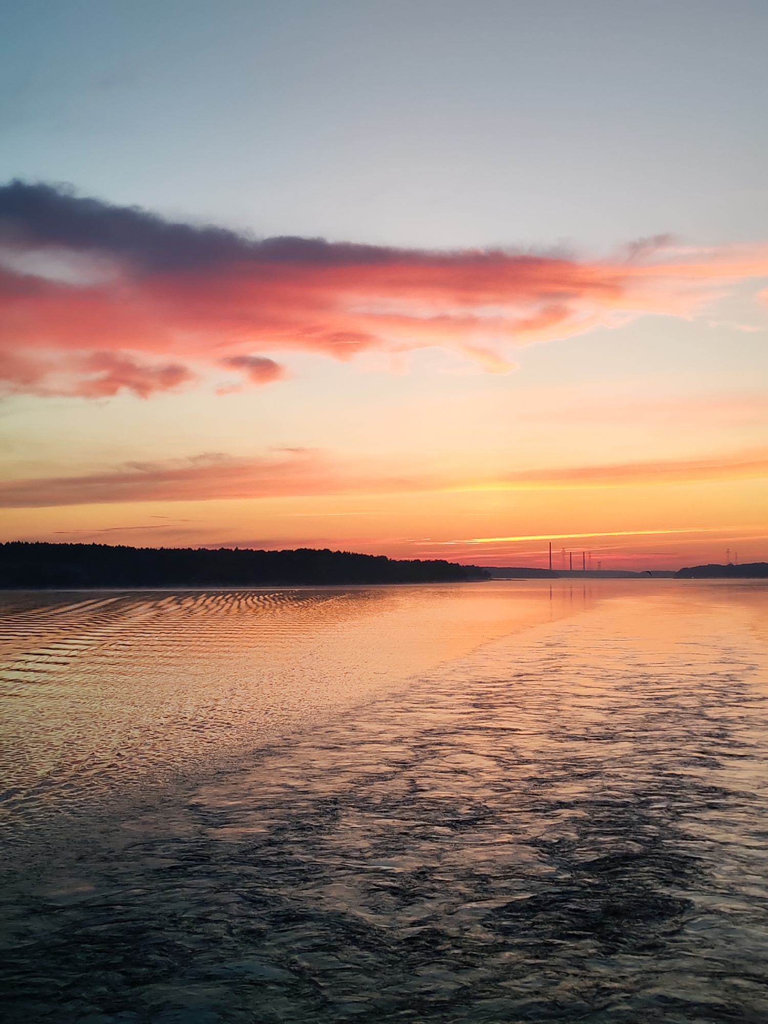 A wide shot captures a serene body of water at sunset, with the sky painted in vibrant hues of orange, pink, and pale blue. Clouds, tinged with pink and purple, drift across the upper portion of the image. The horizon is marked by a dark silhouette of a treeline on the left and distant industrial structures, likely smokestacks, on the right. The water's surface is rippled, reflecting the colorful sky and showing a visible wake trailing towards the viewer, suggesting the image was taken from a moving vessel. The overall impression is one of natural beauty and tranquility at dusk in the vicinity of Плоски, Russia.