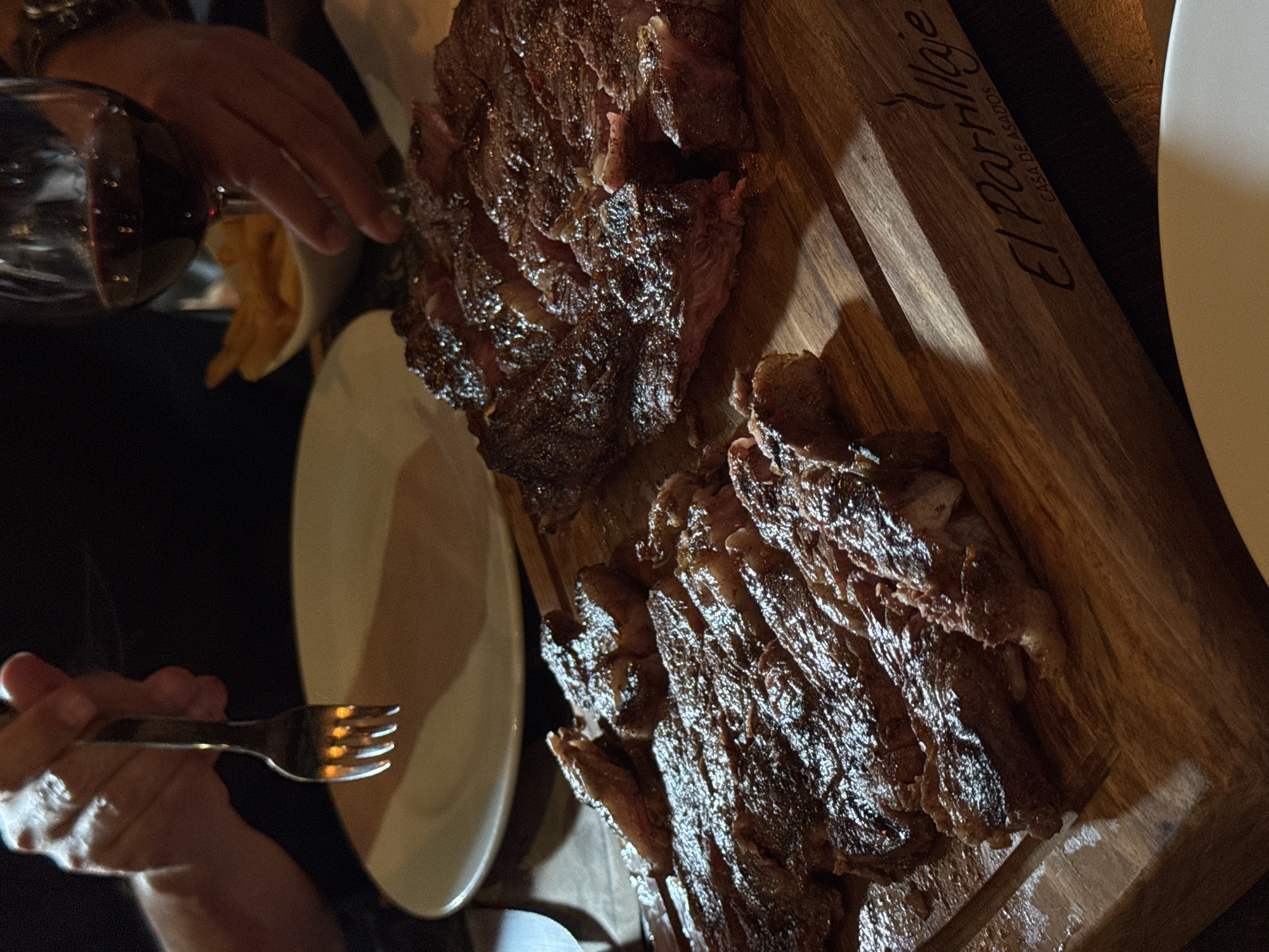A high-angle shot shows a partially eaten, grilled steak on a wooden serving board. The steak is sliced, appearing well-done with dark, caramelized crusts. The board has "El Partilla" and "Casa de Asados" (house of barbecue) printed on its side. A white plate is positioned near the board. A fork is in a person's hand, poised as if to take a piece of the steak. Another person's hand reaches toward the steak from the other side of the table. A wine glass is partially visible in the background. The location is likely a restaurant specializing in grilled meats in Puebla, Mexico, given the Spanish text and the style of cuisine.