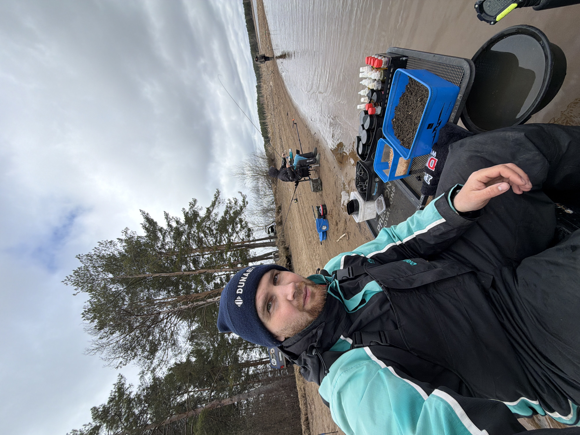 The image shows a man wearing a dark blue beanie with white logos and a black and turquoise jacket. He is positioned on a sandy beach, likely on the shore of a body of water. Behind him, there are several individuals fishing. A blue container filled with dark-colored bait and several containers with colorful liquids are placed next to a black bucket. In the background, there are pine trees and a cloudy sky. The location appears to be Chaykovsky, Russia, based on the environment and the likely angling activity.
