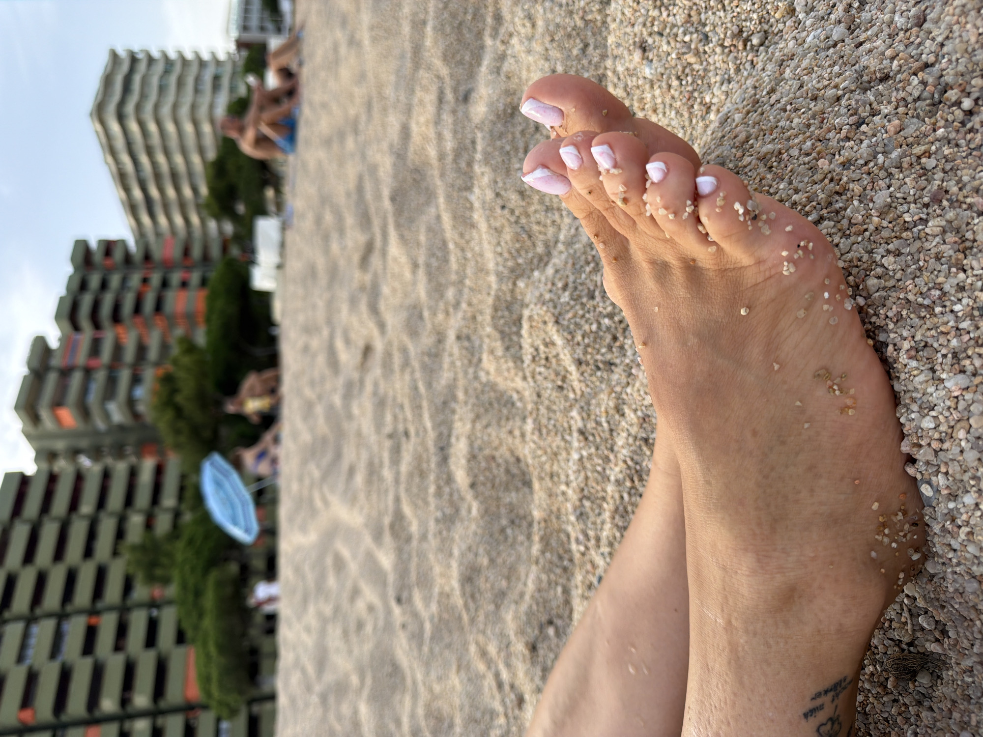 A close-up shot shows a person's feet resting on a pebble beach in Platja D'aro, Spain. The feet are partially submerged in the pebbles, and a tattoo is visible on the left ankle. The toenails are painted with white and light pink French tips. In the background, blurred figures are visible enjoying the beach, with tall, multi-story buildings and trees lining the shore. The sky is clear and bright.