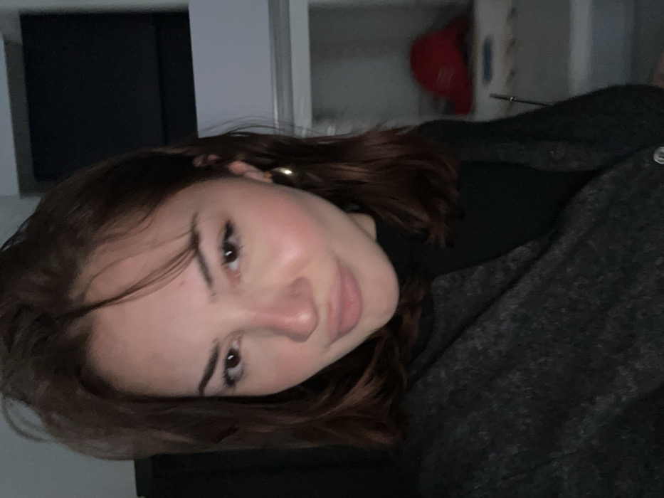 A young woman is featured in a portrait-style photograph taken indoors, rotated 90 degrees clockwise. She has long, dark brown hair that falls around her face. Her expression is soft, with a subtle smirk or gentle smile, and she looks directly at the camera. Notable facial features include well-defined eyebrows and visible eyelashes. A golden hoop earring is discernible on her right ear.
She is wearing a dark-colored top, possibly a black turtleneck, underneath a dark grey, textured jacket or coat. A single silver-toned button or stud is visible on the left side of the jacket. The background is dimly lit, suggesting an evening or night setting indoors, or a room with low artificial light. A red baseball cap hangs on a light-colored surface or wall in the upper right background. Other background elements are indistinct due to the low light. No specific location cues are visible within the image, though the context indicates the location is Strasbourg, France. There is no visible text.