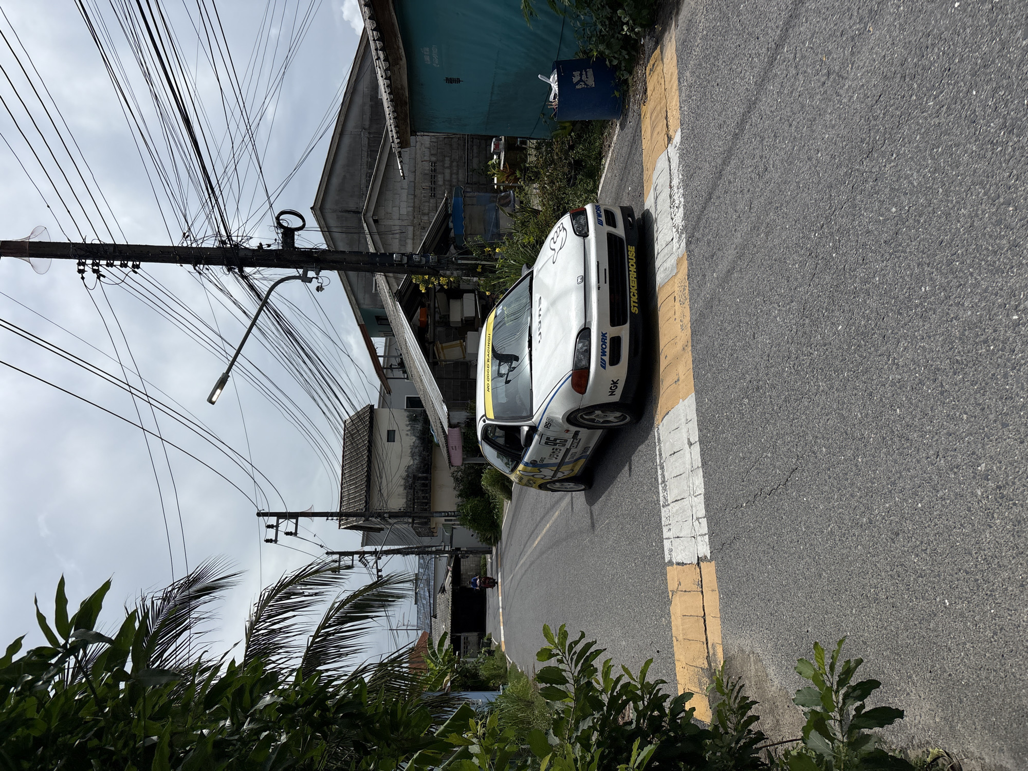A white, modified Honda Civic is positioned on an asphalt road in Choeng Thale, Thailand. The car features racing decals, including the number "95" and "STICKERHOUSE". In the background, a motorcycle is visible in the distance, and various residential buildings with tiled roofs line the street. Overhead, a dense network of utility wires crisscrosses the sky, supported by wooden utility poles. Green foliage from trees and palm fronds frames the left side of the image. The road has a pedestrian crossing with yellow and white markings. A blue trash bin is situated to the right of the car. The sky is overcast with clouds.