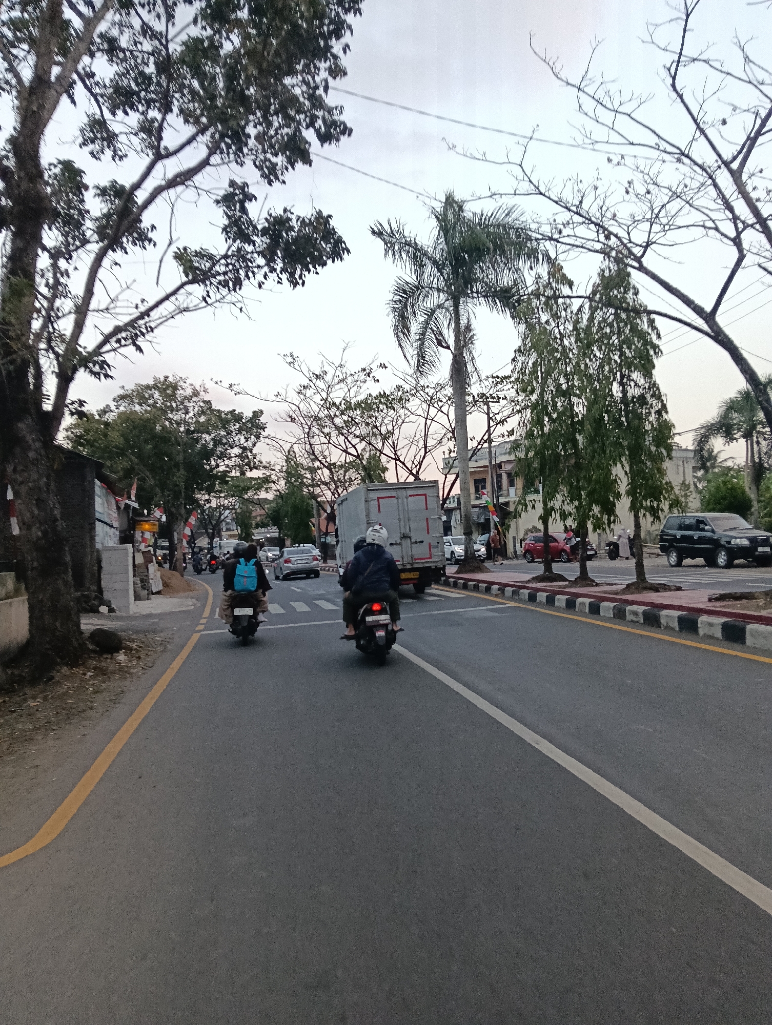 The image depicts a street scene in Tasikmalaya, Indonesia. The road is paved and features yellow and white lane markings. Several vehicles are present, including two motorcycles in the foreground, a large truck, cars, and a black SUV. Two individuals are riding on the motorcycles in the foreground, one wearing a blue backpack. Trees line the sides of the road, with some appearing bare and others with foliage. Buildings and other structures are visible in the background. The sky is overcast, suggesting it may be daytime. The scene appears to be a typical urban street with moderate traffic.