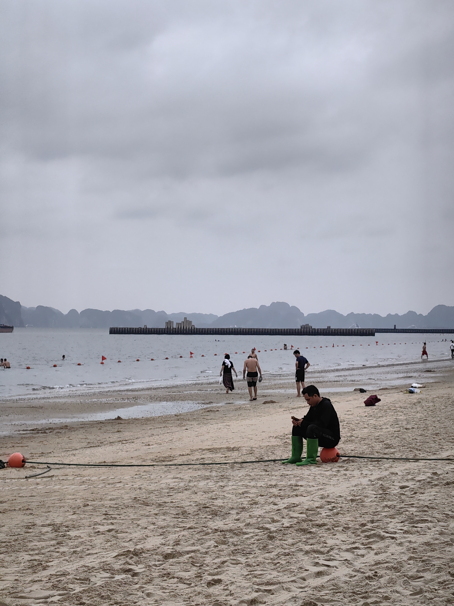 This image captures a tranquil beach scene under an overcast sky in Bai Chay Ward, Vietnam. The overall mood is calm and muted due to the cloudy weather, which diffuses the daylight evenly across the landscape.
In the foreground, a man sits on a bright orange buoy placed directly on the sandy beach. He is dressed in dark trousers and a black long-sleeved shirt, contrasting sharply with his vibrant green rubber boots. He is looking down intently at a mobile phone he holds in his hands. A dark rope runs from the orange buoy further into the foreground. The sand shows various footprints and ripples from receding waves.
The midground features the calm, grey-blue water of the bay, where several people are seen. Some individuals are waist-deep in the water, presumably swimming or wading, while others walk along the water's edge on the wet sand. A line of small orange buoys and red flags marks a designated swimming area in the water.
In the background, the distinctive, rugged silhouettes of limestone karsts (mountains) rise from the water, characteristic of the Ha Long Bay region. Stretching across the horizon in the water is a long, dark man-made structure, possibly a breakwater or pier, with what appears to be more substantial constructions built upon it in the center. Further to the left, a large ship or cargo vessel is visible on the water.
No legible text is visible in the image. The scene suggests a leisure activity on a cooler, cloudy day at the beach.
