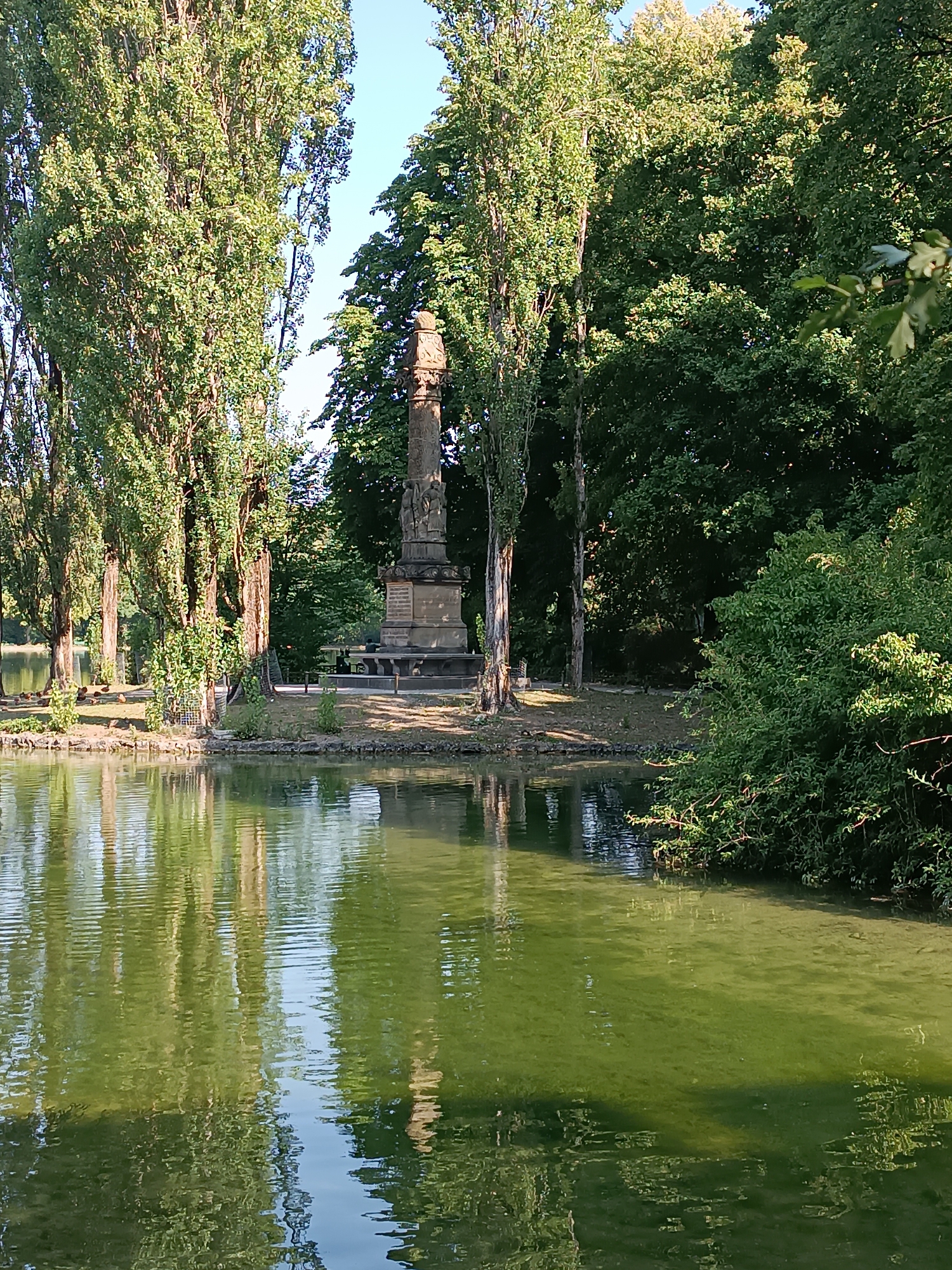 The image shows a monument situated on the edge of a body of water, likely a pond or small lake, surrounded by trees. The monument is a tall, ornate stone structure, possibly a column or obelisk, with carvings or reliefs visible on its upper sections. It appears to be ancient or historical. Several tall, slender trees with dense foliage flank the monument, creating a sense of height and enclosure. The water in the foreground reflects the trees and the monument, creating a distorted but recognizable mirror image on its surface, which is a vibrant green. The surrounding vegetation is lush and green, indicating a park or garden setting. The overall atmosphere is serene and natural. This monument is the "Mariensäule" or Marian Column in the center of Munich's Marienplatz, a prominent public square.