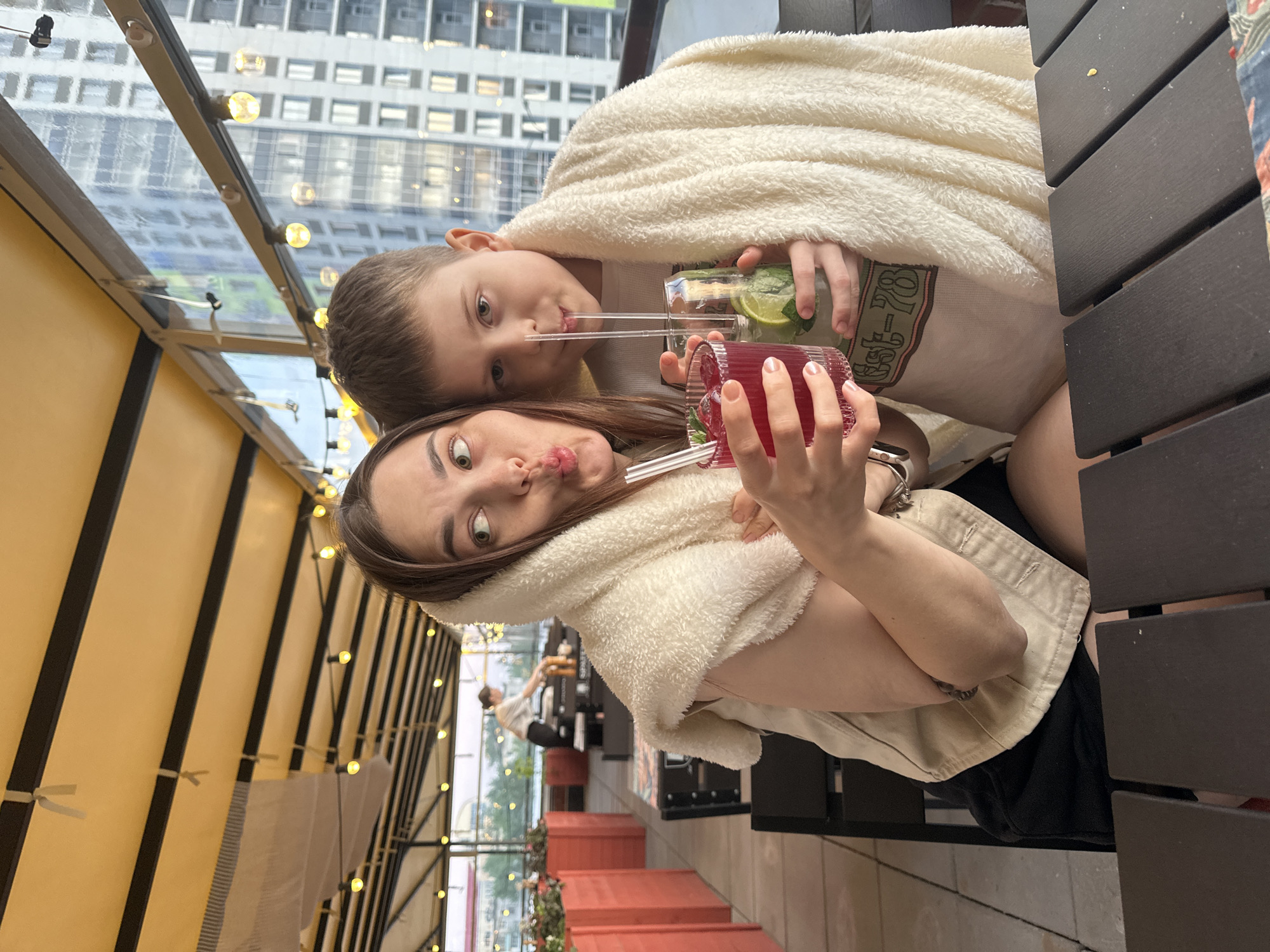 Two people, a woman and a boy, are drinking beverages at an outdoor cafe. The woman is holding a pink drink with a straw and making a face at the camera. The boy is holding a green drink with a straw and looking at the camera. Both are covered in a white, fluffy blanket. They are sitting at a dark wood table. The cafe has a transparent roof and windows that show a city skyline. The cafe is decorated with string lights. The photo was taken in Perm, Russia. There are red flower boxes with greenery along the cafe's perimeter. Another person is sitting in the background.