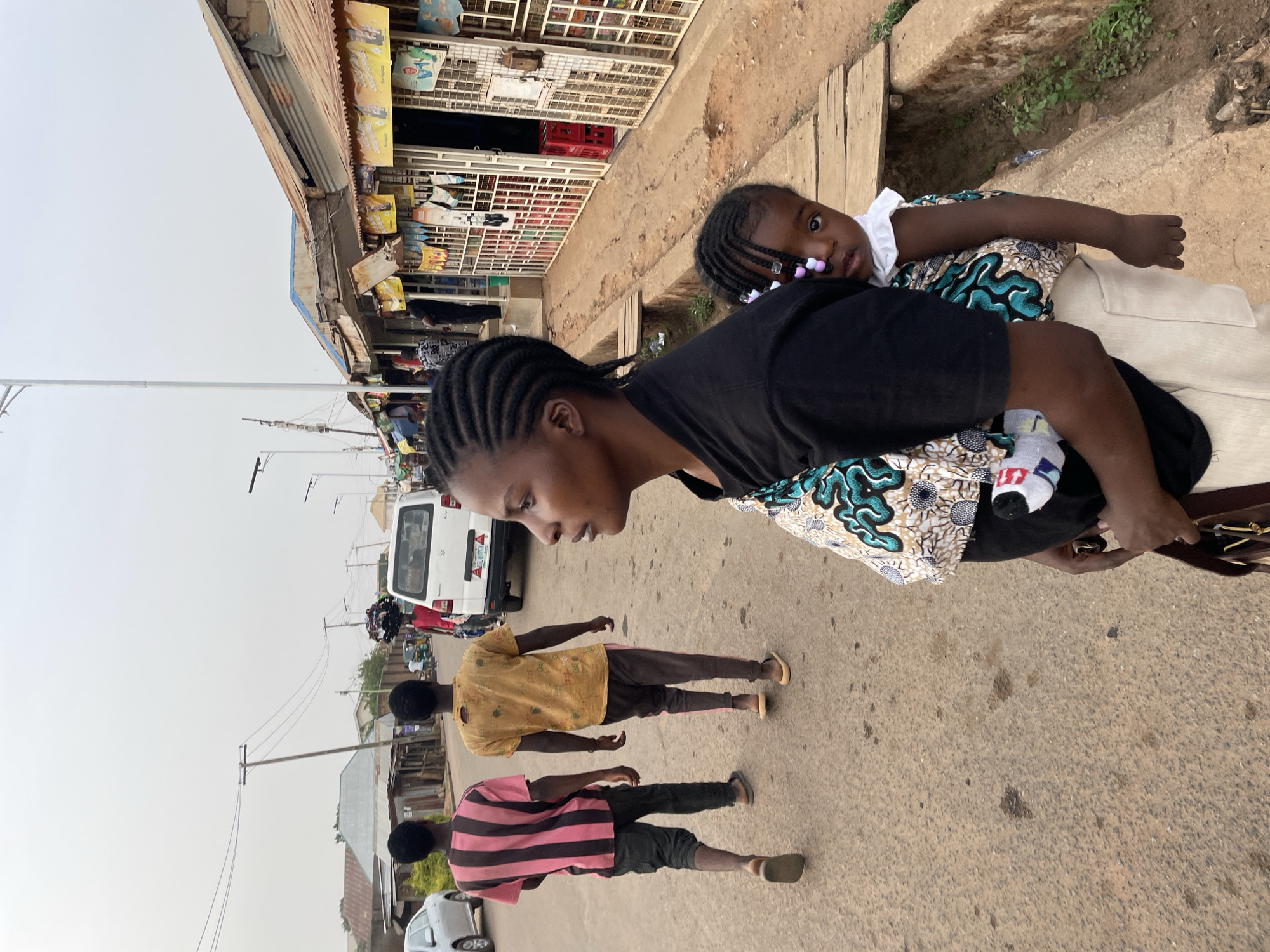 This media file captures a vibrant street scene in Jos, Nigeria, during the daytime under what appears to be an overcast or hazy sky. The primary subjects are a woman, positioned prominently in the right foreground, and a young child carried on her back in a traditional fabric wrap. The woman has braided hair and wears a black top over a patterned fabric. The child, also with braided hair adorned with pink and white beads, wears a white top and patterned clothing, looking directly towards the viewer with a neutral expression. One of the child's feet, visible, shows a mismatched pair of socks.
In the mid-ground, two men are walking away from the camera. One wears a yellow shirt and dark pants, and the other a pink and black striped shirt and dark pants, both wearing sandals. The street they walk on is a light-colored asphalt or concrete, showing some wear.
The left side of the frame is lined with single-story commercial buildings or market stalls, characterized by corrugated metal roofs and metal grate fronts, indicating a bustling commercial area. Various items and products are visible within the stalls, though specific details are blurry. A white van or bus is parked further down the street in the background, and utility poles with overhead wires are visible, extending into the distance. The overall atmosphere suggests a typical daily scene in an urban or semi-urban setting. No clear, legible text is visible on any signs or vehicles.