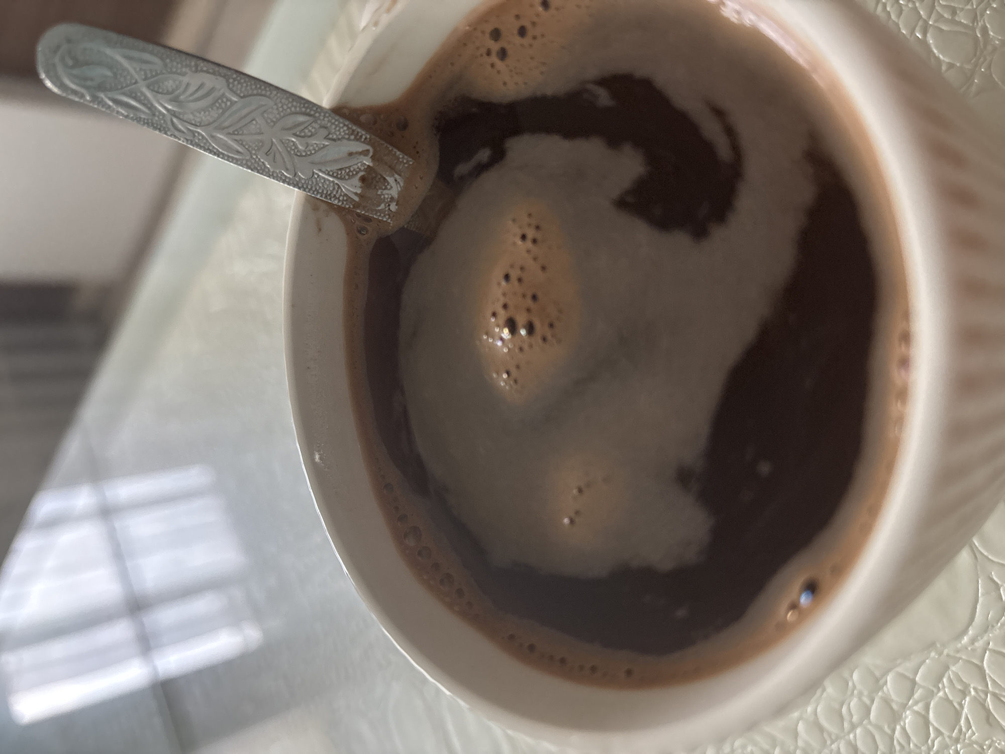 A high-angle, close-up shot shows a white ceramic mug filled with a dark brown beverage, possibly coffee with milk. A silver-colored spoon with decorative etchings is partially submerged in the liquid. The mug sits on a white surface with a textured, slightly reflective pattern. Window light with a visible frame is reflected on the surrounding surface. The city of Chaikovsky, Russia is not discernible from the image.