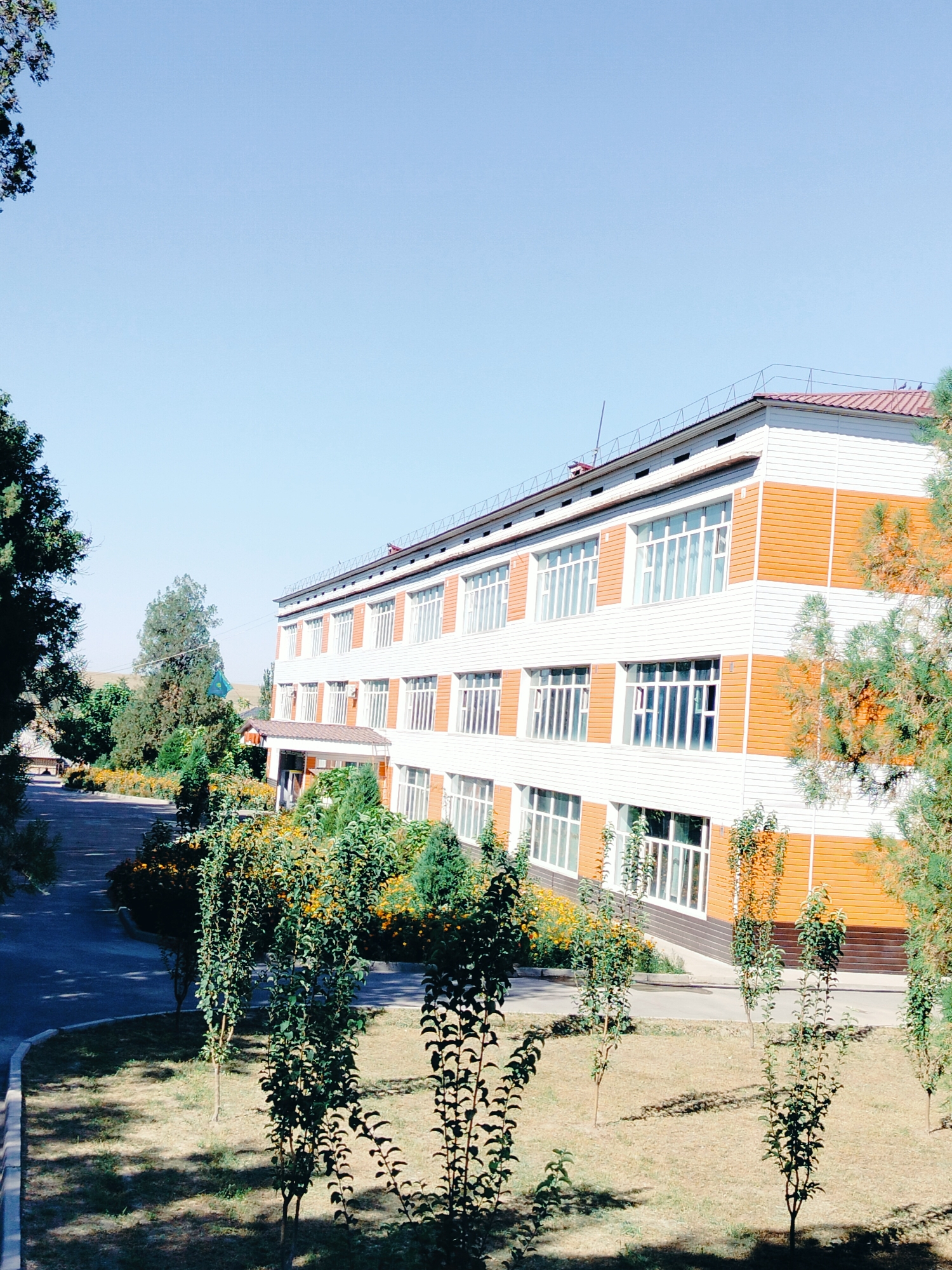 The image shows a modern, multi-story building with white and orange siding, and numerous windows. The building appears to be a school or educational institution, with a fenced rooftop. In front of the building, there is a paved walkway or road, a grassy lawn with trees and yellow flowers, and some bushes. The sky is clear and blue. The scene is set in an outdoor environment, likely in a suburban or rural area given the presence of open land and trees. The specific location is identified as the city of Kölkent, Kazakhstan, suggesting it's a public building within that region. There are no people visible in the image.