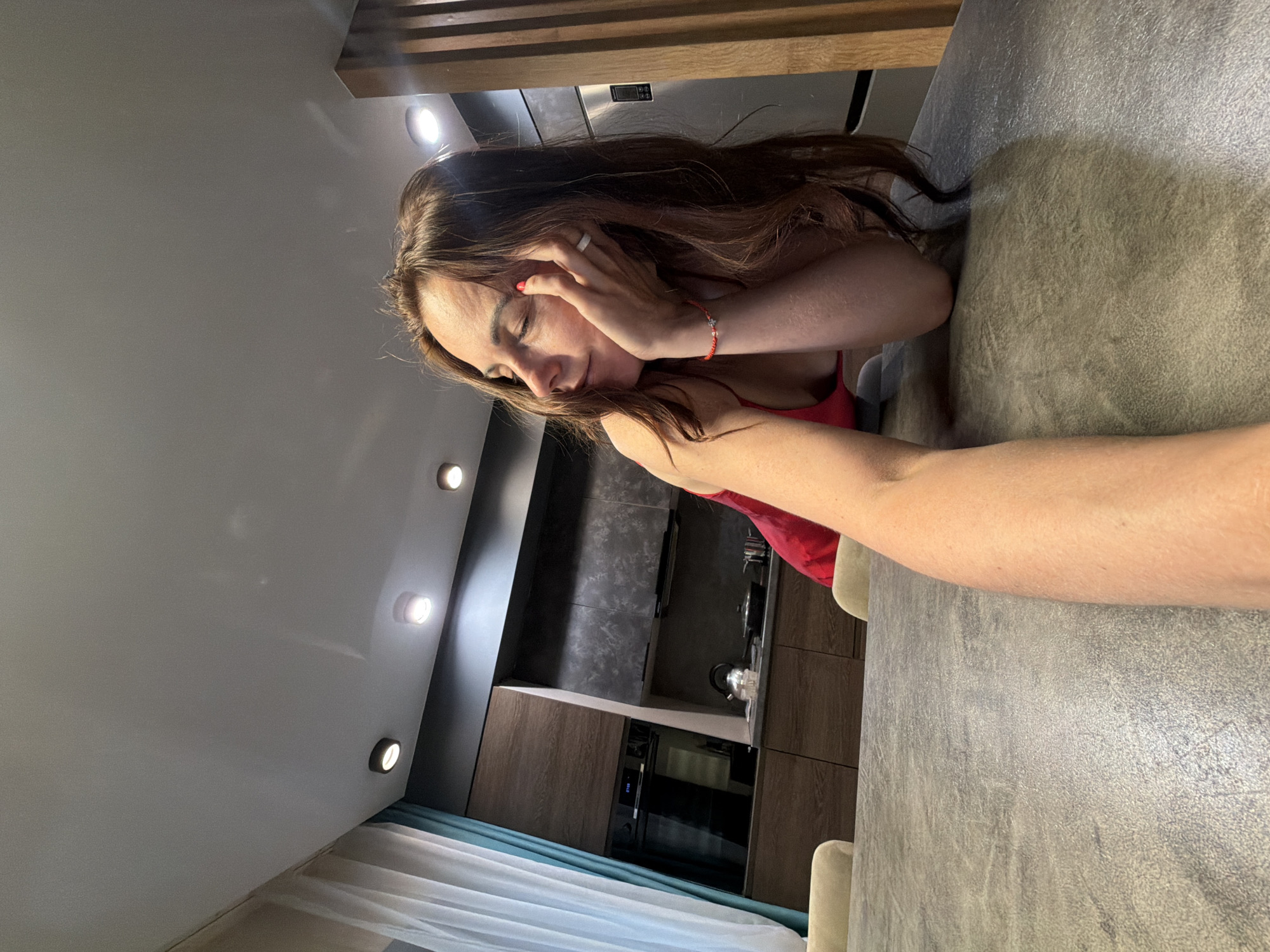 A woman with long, dark brown hair, fair skin, and wearing a red top, is depicted in a domestic setting. She is seen with her eyes closed and her left hand gently touching her face. Her fingers display red nail polish, and she wears a white ring on her left ring finger and a red string bracelet with a small charm on her left wrist. Her right arm extends into the foreground, suggesting a selfie or close-up shot.
She is leaning on a large, textured grey table or counter. The background reveals a modern kitchen area with dark wooden cabinets, a dark grey tiled backsplash, and built-in appliances, including an oven or microwave with an illuminated but illegible digital display. A metallic kettle is visible on the kitchen counter. The ceiling is fitted with multiple recessed circular lights, casting a soft glow. Light blue and white curtains or blinds are visible on the left side of the frame, and some wooden slats appear at the top.
The woman appears to be in a relaxed state, possibly resting or in quiet contemplation. The overall atmosphere of the room is contemporary and cozy, illuminated by artificial lighting, which might indicate evening or nighttime. The location context for this scene is Birobidzhan, Russia. No legible text is visible in the image.