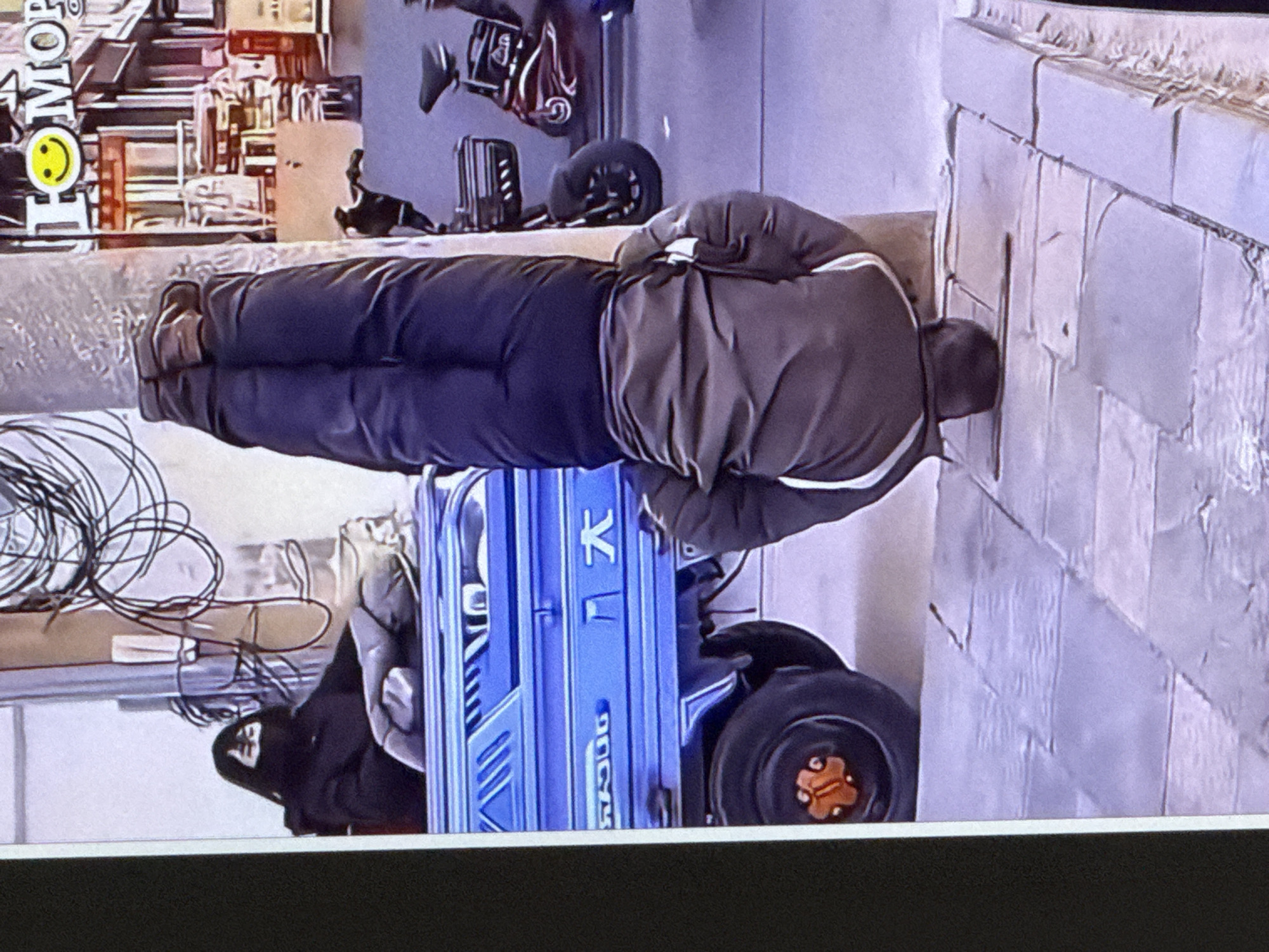 A man is performing a handstand with his feet on a pole in the city of Cahul, Moldova. He is wearing dark pants and a brown jacket. To his left, a woman wearing a mask and dark clothing is sitting in a blue truck. In the background, there are buildings and two parked electric scooters. The ground is paved with stone tiles.