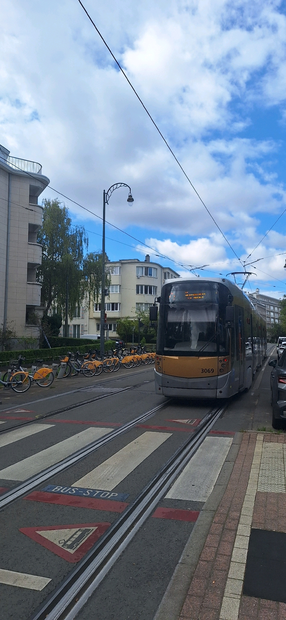 In the city of Ixelles - Elsene, Belgium, a modern, silver and gold tram numbered 3069 is positioned on tram tracks, facing forward. The tram has its headlights illuminated. Adjacent to the tram tracks, a row of yellow rental bicycles are parked neatly. The scene is set on a street with a pedestrian crossing marked by white stripes and red lines. A "BUS-STOP" sign is visible on the road surface, along with a triangular warning sign depicting a tram. On the left side of the street, a multi-story building with curved balconies and large windows stands tall, partially obscured by a weeping willow tree. Across the street, another building with a similar architectural style and multiple windows is visible. The sky is partly cloudy, with a clear blue sky visible through scattered white clouds. The overall environment appears to be an urban setting with public transportation infrastructure.
