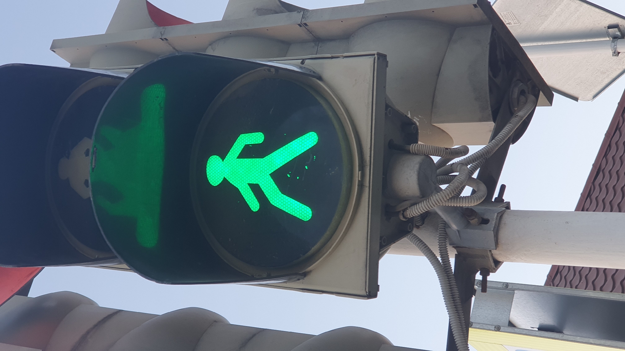 The image shows a close-up of a pedestrian traffic signal displaying a green walking person symbol. The signal is mounted on a metal pole with various wires and conduits attached. Above the green pedestrian signal, a traffic signal housing is visible, and a red triangular sign is partially seen at the top left. In the background, a clear sky and a tiled roof of a building are present, suggesting an outdoor urban environment. The image does not contain any people or specific landmarks that would identify the exact location within Ust-Labinsk, Russia. The lighting suggests it is daytime, possibly late afternoon or early morning due to the angle of the light.