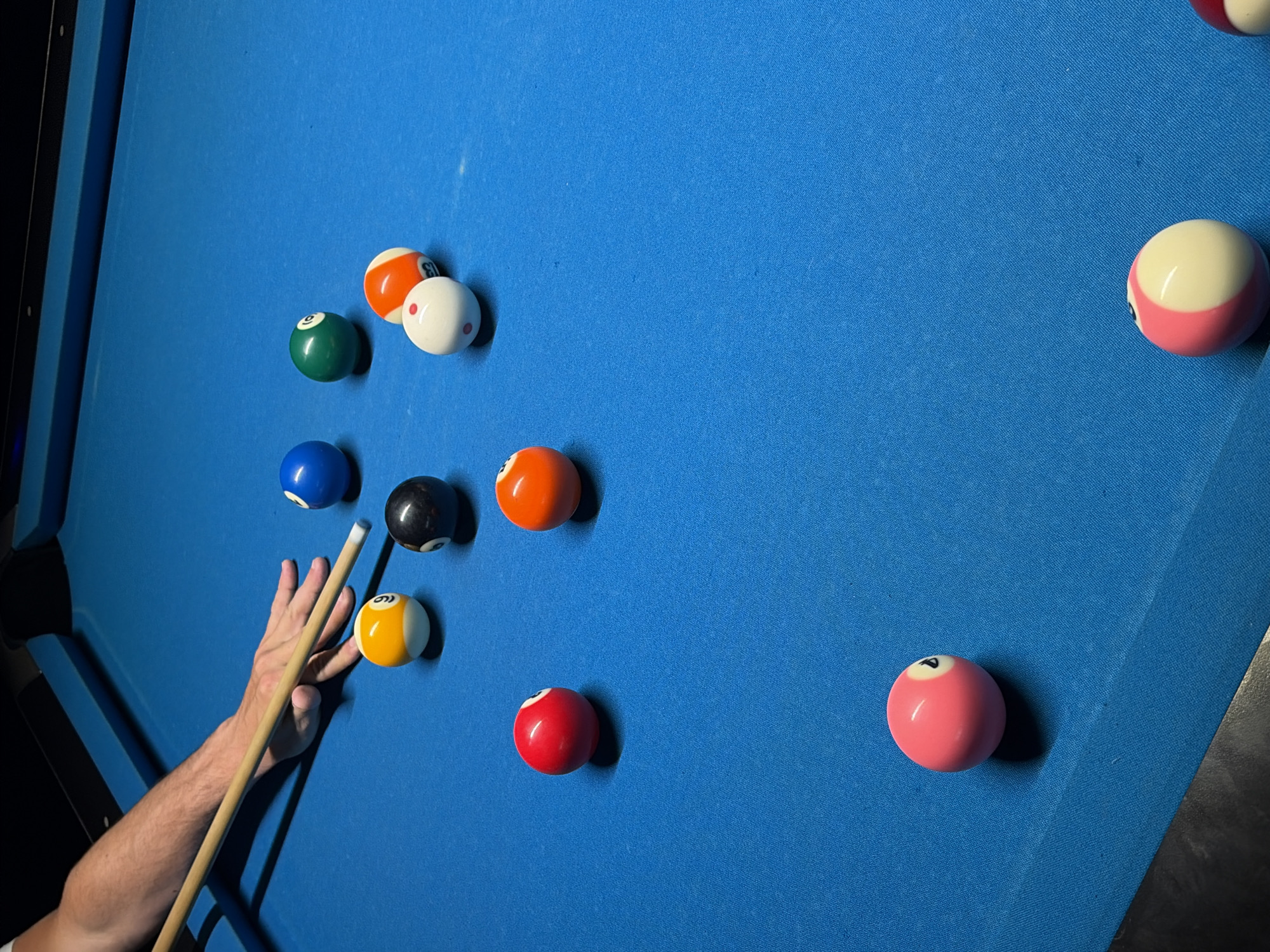 The image captures a close-up, elevated view of a person aiming a pool cue at a yellow 9-ball on a blue felt pool table in an indoor setting, location Unknown, Unknown.
A person's light-skinned forearm and hand are visible in the upper left, with the hand forming a bridge on the blue felt. The person holds a light-colored wooden cue stick, poised to strike the yellow 9-ball, which is positioned centrally in the upper left quadrant. To the immediate right of the 9-ball is a black 8-ball.
Eleven pool balls are distributed across the blue felt surface. In the upper central and right areas, from left to right, are a solid blue 2-ball, a solid green 6-ball, a solid red 3-ball, a striped orange 5-ball, and a solid orange 4-ball. A white cue ball with distinct red dots is positioned near the orange 5-ball. In the lower left quadrant, a solid pink 7-ball is visible. In the lower right quadrant, there is a pink and white striped ball and a red and white striped ball. The pool table has dark, possibly black, rails visible along the top and bottom edges of the frame. The lighting appears to be overhead, casting soft shadows from the balls and cue stick onto the felt. The overall environment suggests a well-lit game room or pool hall.