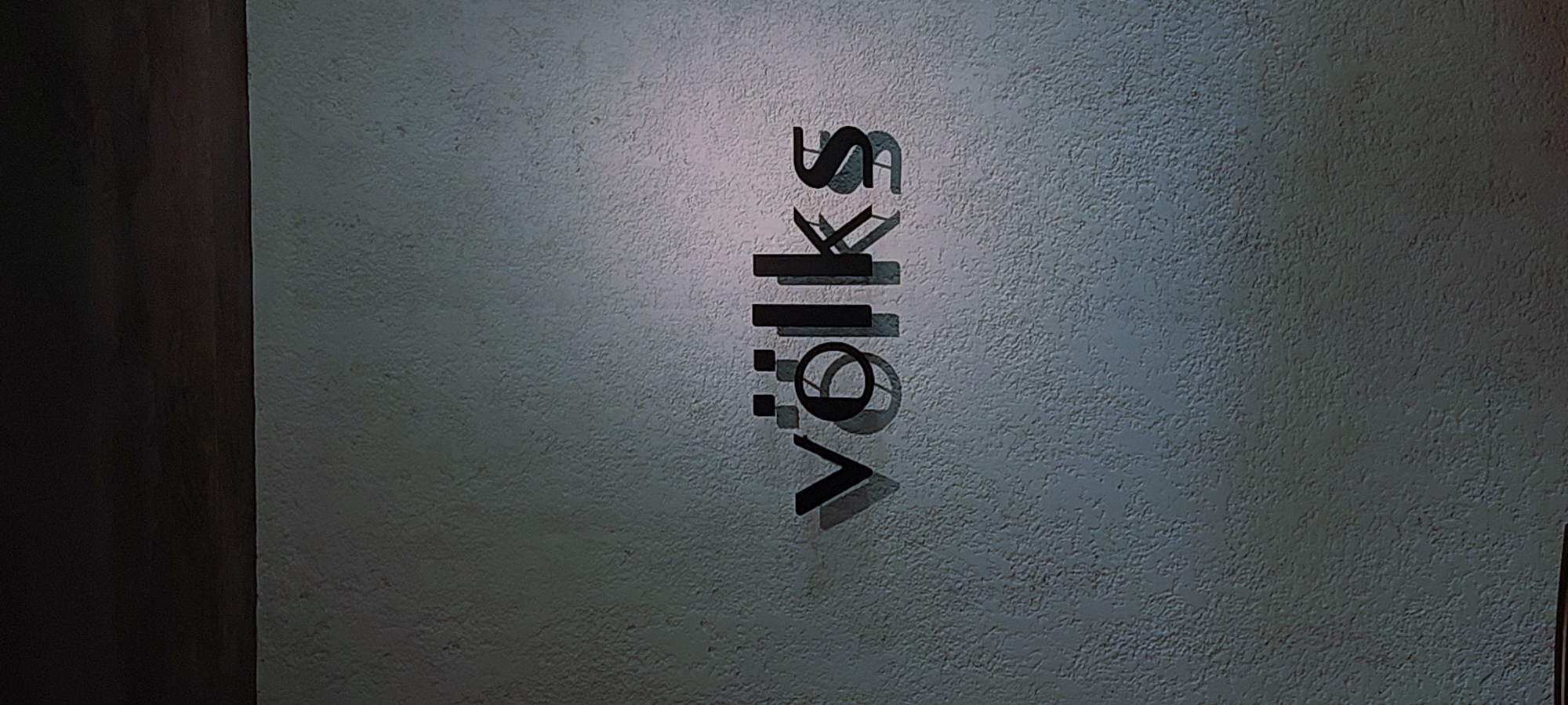The image displays the word "völks" in a stylized, dark font, appearing to be a sign or logo, mounted on a textured, light-colored wall. The letters cast shadows on the wall, indicating a light source from above and to the left. The upper portion of the image shows a dark, horizontal element, possibly a ceiling or a shelf, casting a shadow on the wall below it. The wall has a rough, granular texture. The scene is devoid of people and appears to be an indoor or covered outdoor setting. The location in Cicalengka, Indonesia is not discernible from the visual elements within the image itself.