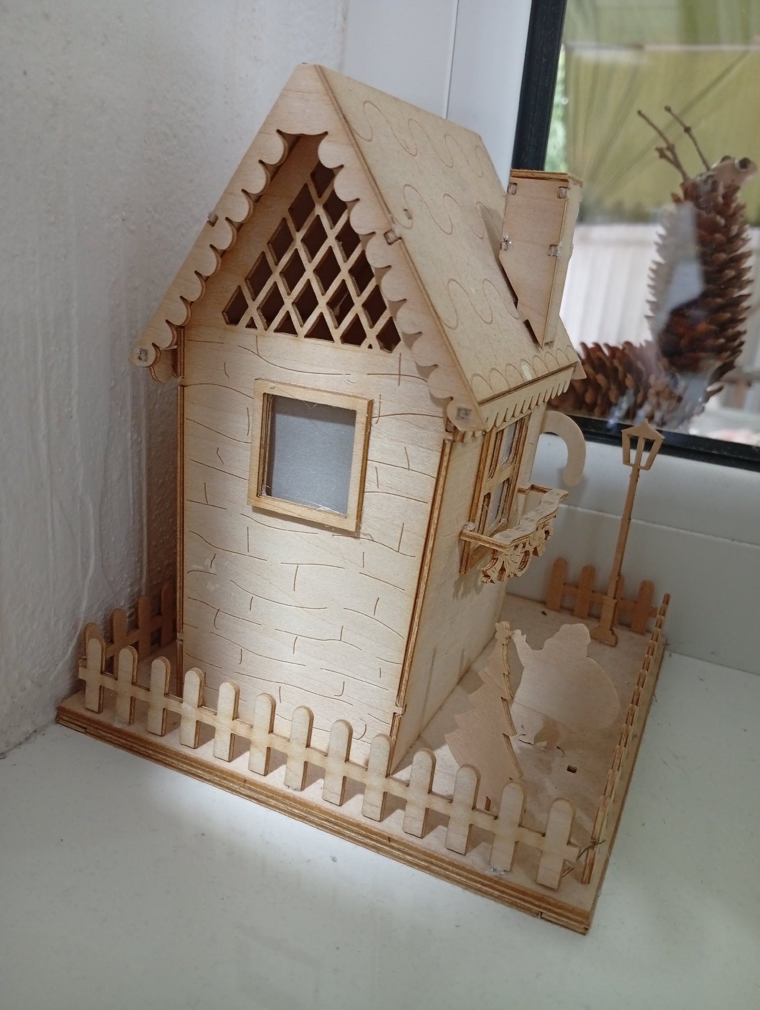 The image shows a small, unfinished wooden model of a house, likely a craft project. The house has a pitched roof with decorative scalloped edges and a diamond-shaped lattice design on one gable end. A small window is visible on the front of the house, and a balcony with intricate wooden detailing extends from a second-story window. A miniature picket fence surrounds the base of the house. Inside the fenced area, two flat, silhouette-style wooden figures are present; one appears to be a person with a large bag. To the right of the house, a tiny street lamp model stands. The model is placed on a white surface, and the background shows a textured white wall on the left and a window with a pine cone visible outside on the right. The scene does not depict any specific location in Pănășești, Moldova, or any other city.