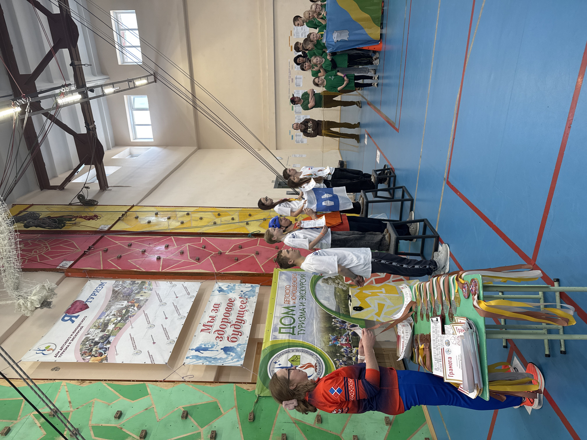 The image shows an indoor gymnasium in Usolye, Russia. A woman wearing a blue and orange athletic outfit stands by a table adorned with medals and certificates labeled "Gramota". A group of children in white shirts stand on black metal stands behind the table. In the background, a group of people in green shirts hold a flag. On the left, a climbing wall has multiple banners affixed, including one that reads "Я туризм", "Мы за здоровое будущее", and "Дом детского и юношеского туризма и экскурсий, г. Березники", referencing the Children's and Youth Tourism and Excursion Center in Berezniki. The gymnasium has a blue floor with red lines and windows near the ceiling. Overhead are industrial metal structures and wires.