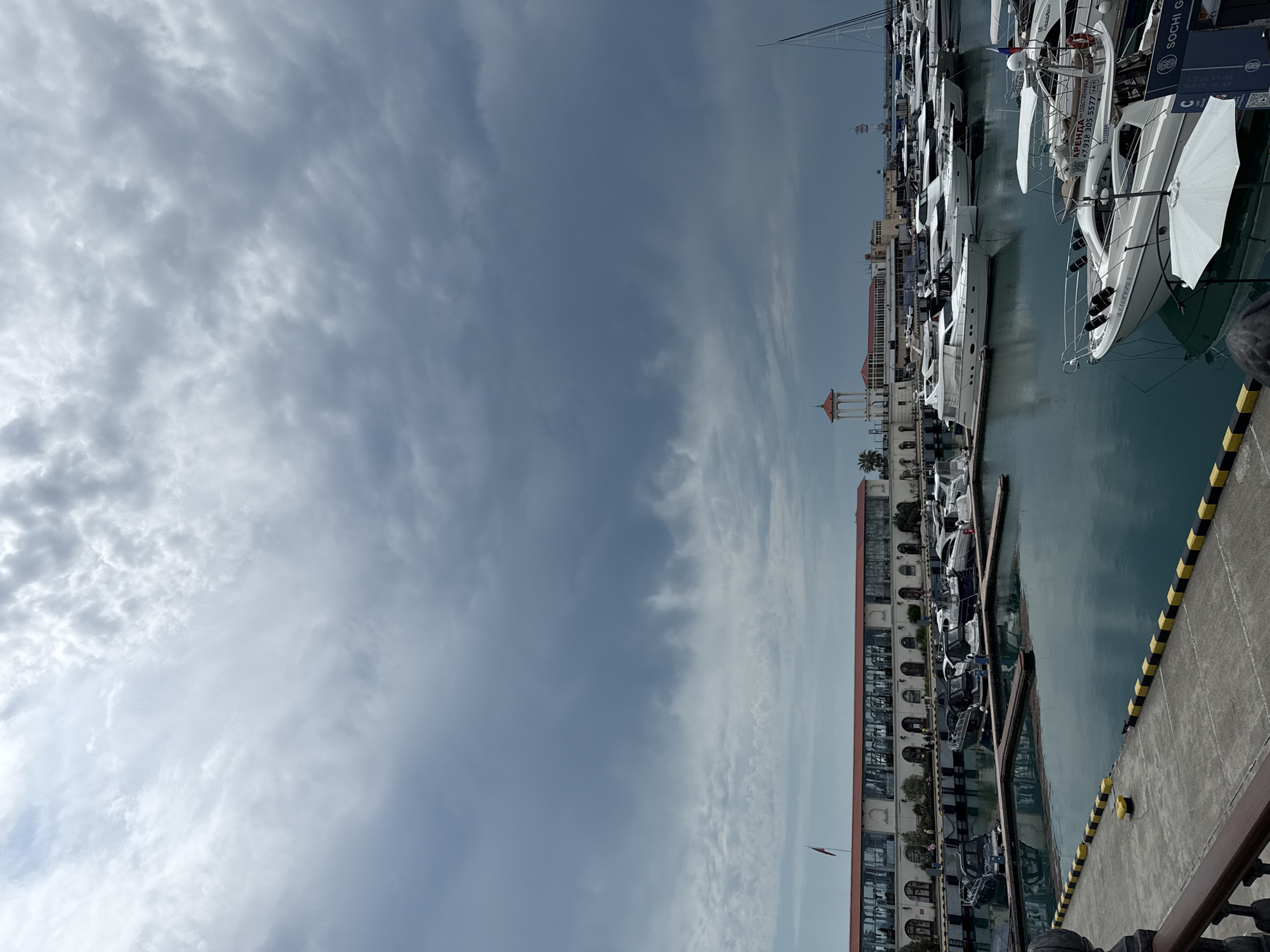 The image captures a view of the Sochi Sea Port under a cloudy sky. Numerous yachts and boats are docked in the harbor, with their reflections visible in the calm, dark blue water. A long, white building with arched windows and a red-tiled roof lines the waterfront, possibly housing shops or restaurants. A flagpole with a red flag stands near the building. In the background, a white tower with a red roof and a tall, slender mast of a sailboat are visible. The foreground shows a concrete pier with yellow and black checkered markings along the edge. The overall atmosphere suggests a serene but possibly impending change in weather, given the heavy cloud cover.