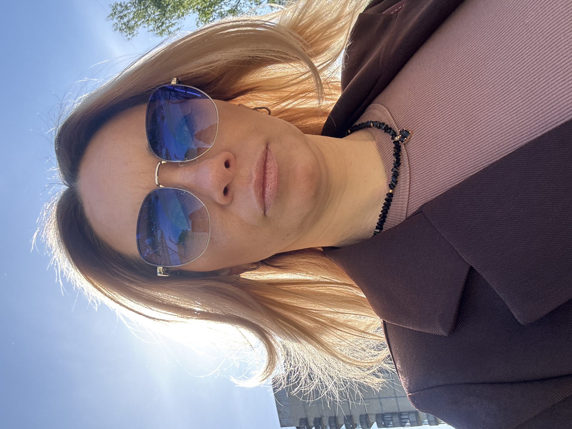 A woman is photographed at a low angle, with a building visible in the background. She wears sunglasses with blue tinted lenses that reflect the surrounding environment, and has blonde hair. She is wearing a lavender top and a dark brown jacket with a collar. A beaded necklace is visible around her neck. The sky is blue. The location is Vsevolozhsk, Russia.