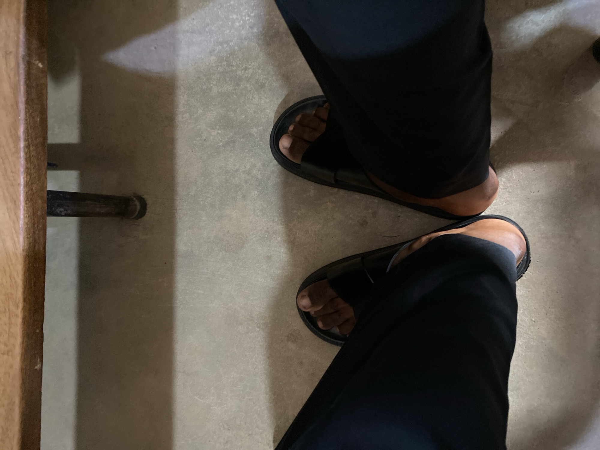 The image shows a person's legs and feet wearing black slide sandals and black pants. The person is sitting under a wooden table with a metal leg visible. The ground appears to be concrete. The lighting suggests an indoor or shaded outdoor environment. The scene is captured from a high-angle, overhead perspective, looking down at the subject's lower body. There are no discernible landmarks that would definitively place the location in Jos, Nigeria, or any other specific city. The image focuses on the immediate area around the person's feet and the furniture.