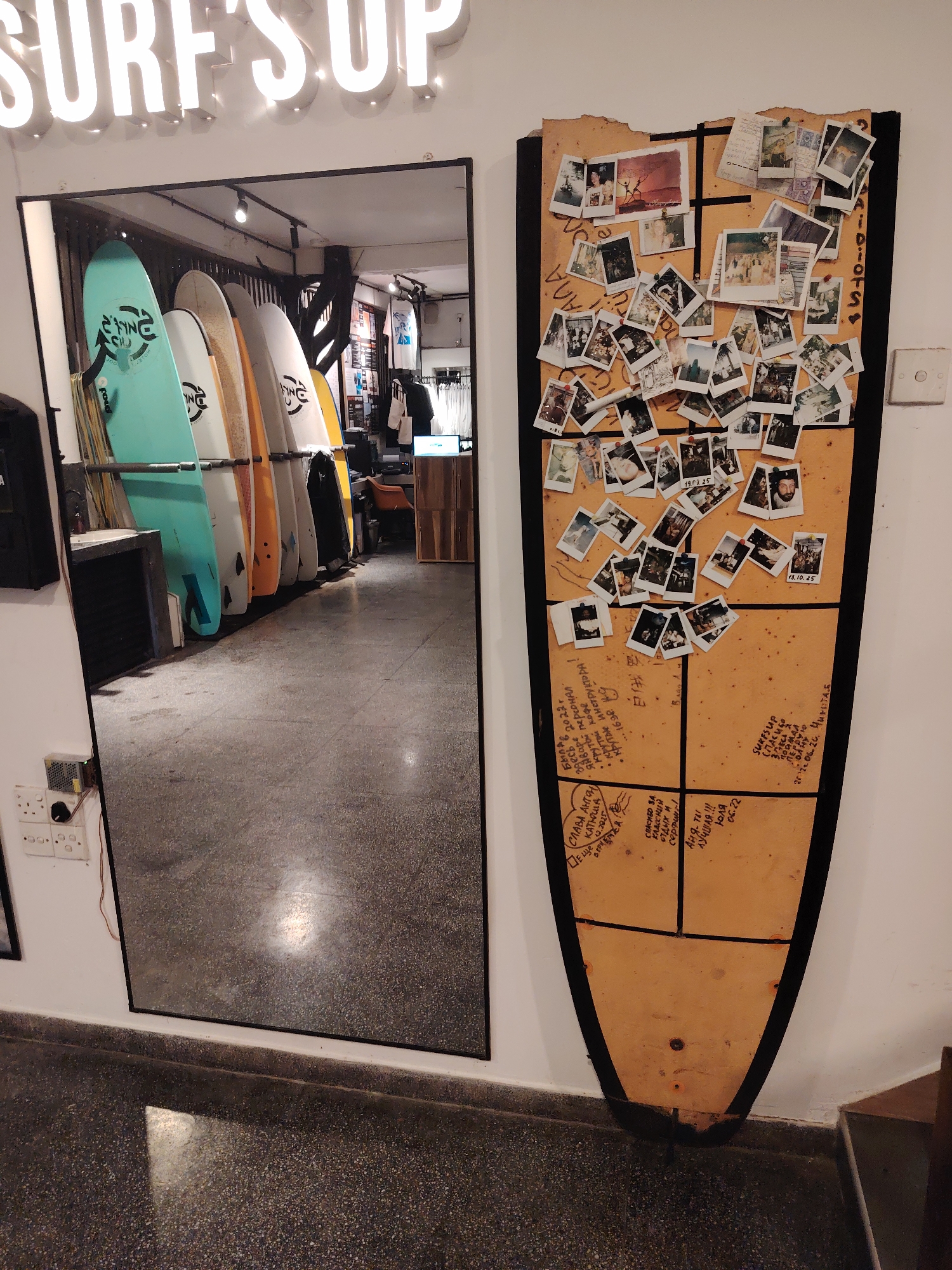 This image captures an indoor commercial or recreational space, possibly a surf shop, cafe, or lounge, in an unspecified city and country. The scene features a large full-length mirror, a surf-themed display board, and a neon sign.
On the upper central portion of the wall, a neon sign spells out "SURF'S UP" in illuminated white capital letters. Below this, a large, black-framed rectangular mirror occupies the left side of the frame, extending from mid-wall to the floor. The mirror reflects a row of at least six surfboards of varying sizes and colors (teal, white, orange, yellow, and black), standing upright in a rack. Reflected beyond the surfboards are elements of a retail interior, including clothing racks, a light-colored counter, and overhead spotlights. On the white wall immediately to the left of the mirror, a white electrical outlet with multiple sockets is mounted, beneath which a small electronic component with a green circuit board and fan, possibly a power supply unit, is attached.
To the right of the mirror, a large, vertically oriented display board shaped like a surfboard is mounted on the white wall. The board is made of a light-colored wood, possibly plywood, with a black outline and is horizontally segmented into four main quadrants in its lower half. The upper portion of the board features a roughly torn, jagged edge. Numerous small, square-format instant photographs, depicting people, social gatherings, and other scenes, are pinned to the upper two-thirds of the board, some overlapping. Several photos have dates handwritten on them, such as "19.01.25" and "13.10.25." Handwritten text, predominantly in Cyrillic (Russian) and some English, covers various sections of the wooden board, both around the photos and in the lower quadrants. Examples of discernible text include "AIIDIOIS" or "3 IDIOTS" vertically along the right edge, and messages of gratitude and well-wishes with names and dates in the lower sections (e.g., "СЛАВА АНТОН КАТЮША 06.2023", "АНЯ ТЫ ЛУЧШАЯ!!! ЮЛЯ 06.22", "SHKESSP СПАСИБО ЗА ПОМОЩЬ").
The floor throughout the visible area is a dark grey speckled terrazzo or concrete, showing some scuff marks and a light reflection near the mirror's base. No individuals are directly present in the foreground; people are only depicted within the small photographs on the display board. The walls are uniformly white.