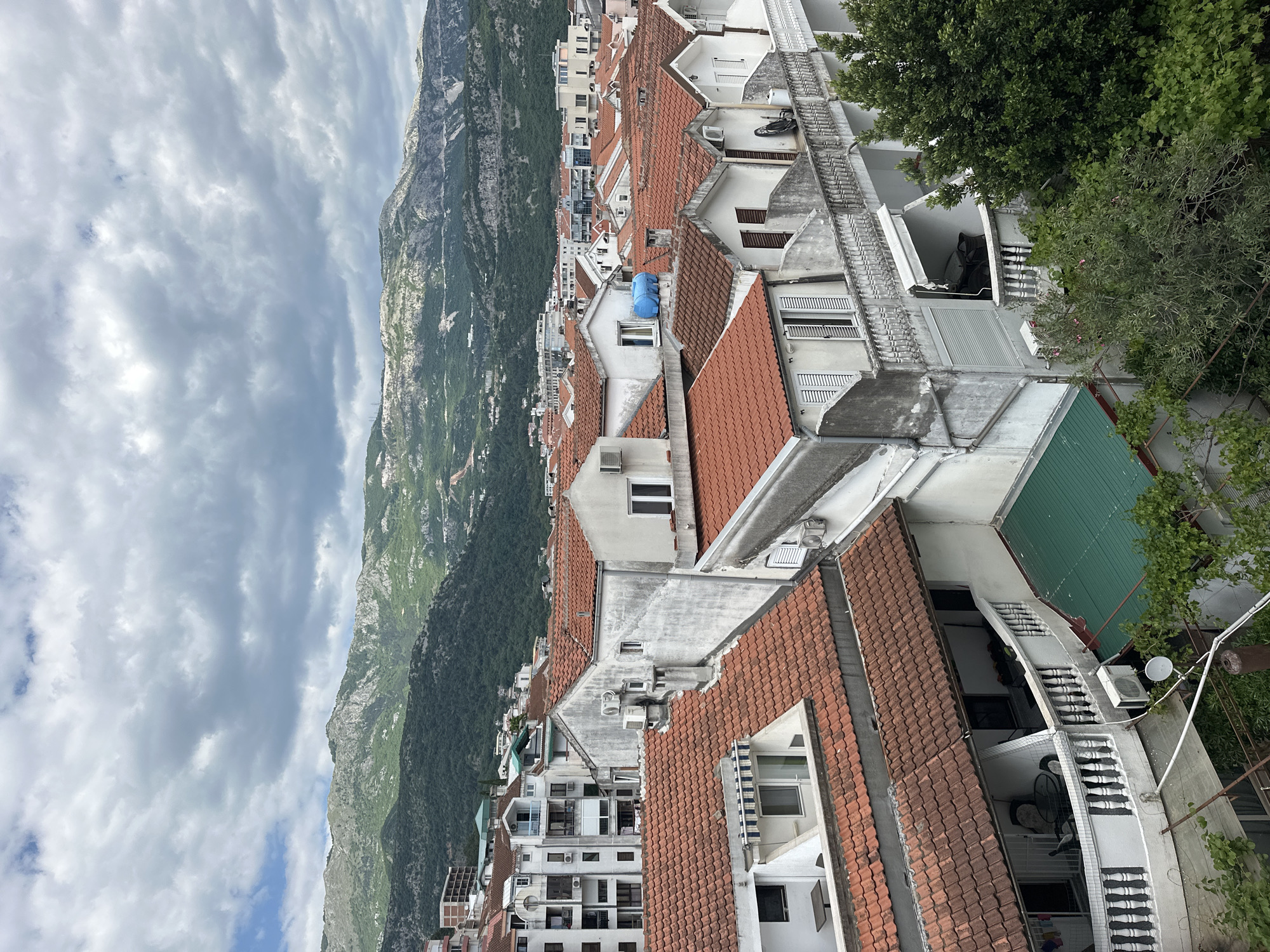 The image captures a high-angle, slightly tilted view of Budva, Montenegro, showcasing a dense urban landscape nestled against a backdrop of rugged mountains and a cloudy sky. The foreground is dominated by the terra cotta tiled roofs of residential buildings. Many buildings are white with dark-shuttered windows. The buildings are closely packed, with balconies and courtyards visible. Air conditioning units are attached to the exterior walls. In the middle ground, mountains rise sharply, partially obscured by clouds. The sky is overcast. There are trees in the scene.