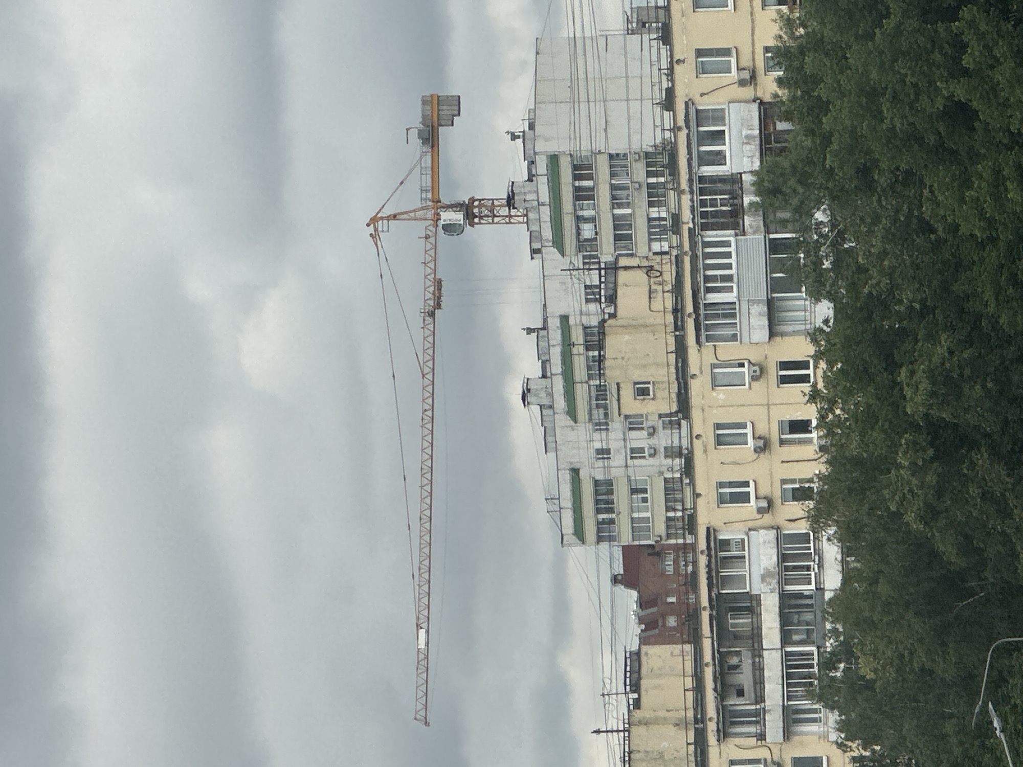 A construction crane with an orange boom and mast extends horizontally across a cloudy, overcast sky above a row of apartment buildings in Moscow, Russia. The buildings appear to be a mix of Soviet-era architecture, with some sections featuring yellow facades and balconies, and others a lighter, possibly white or light grey, color. Green-roofed sections are visible on some of the taller buildings. The lower portion of the image is filled with a dense canopy of green trees. A lamppost is partially visible in the lower left corner.