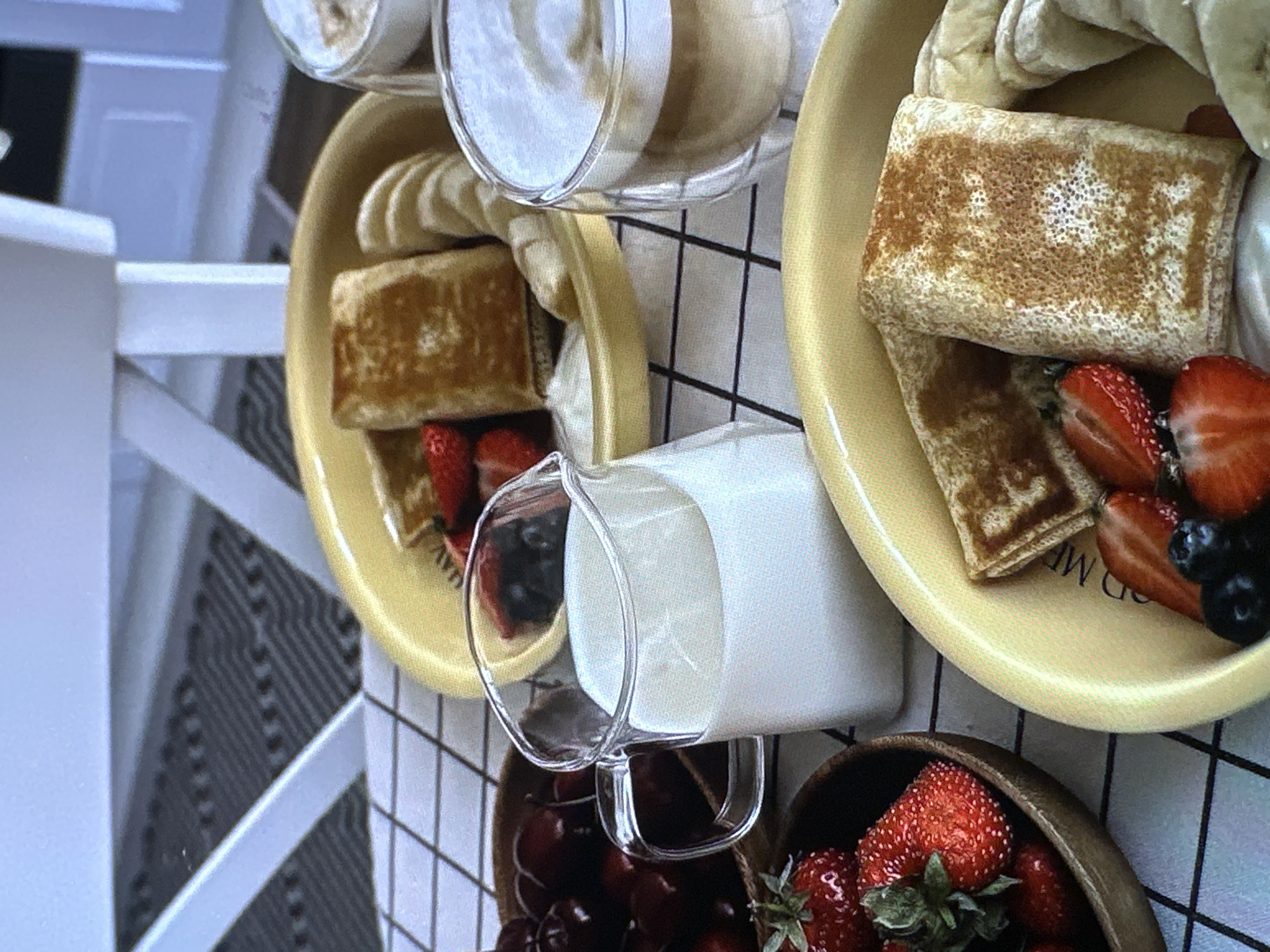 The image shows a tabletop scene with breakfast or brunch items. Two yellow plates are visible, each containing what appears to be folded crêpes or pancakes, banana slices, and a mix of fresh berries including strawberries and blueberries. A small glass pitcher filled with milk rests on the checkered tablecloth, with the bottom edge of the pitcher sitting above wooden bowls filled with more berries, including cherries and strawberries. A glass cup with a white liquid (possibly yogurt or a smoothie) is situated near the plates. The scene is set on a black and white checkered surface that could be a tablecloth or tiles. Part of a white chair can be seen on the left side of the frame. The location and city of Yerevan, Armenia cannot be determined from this image alone.