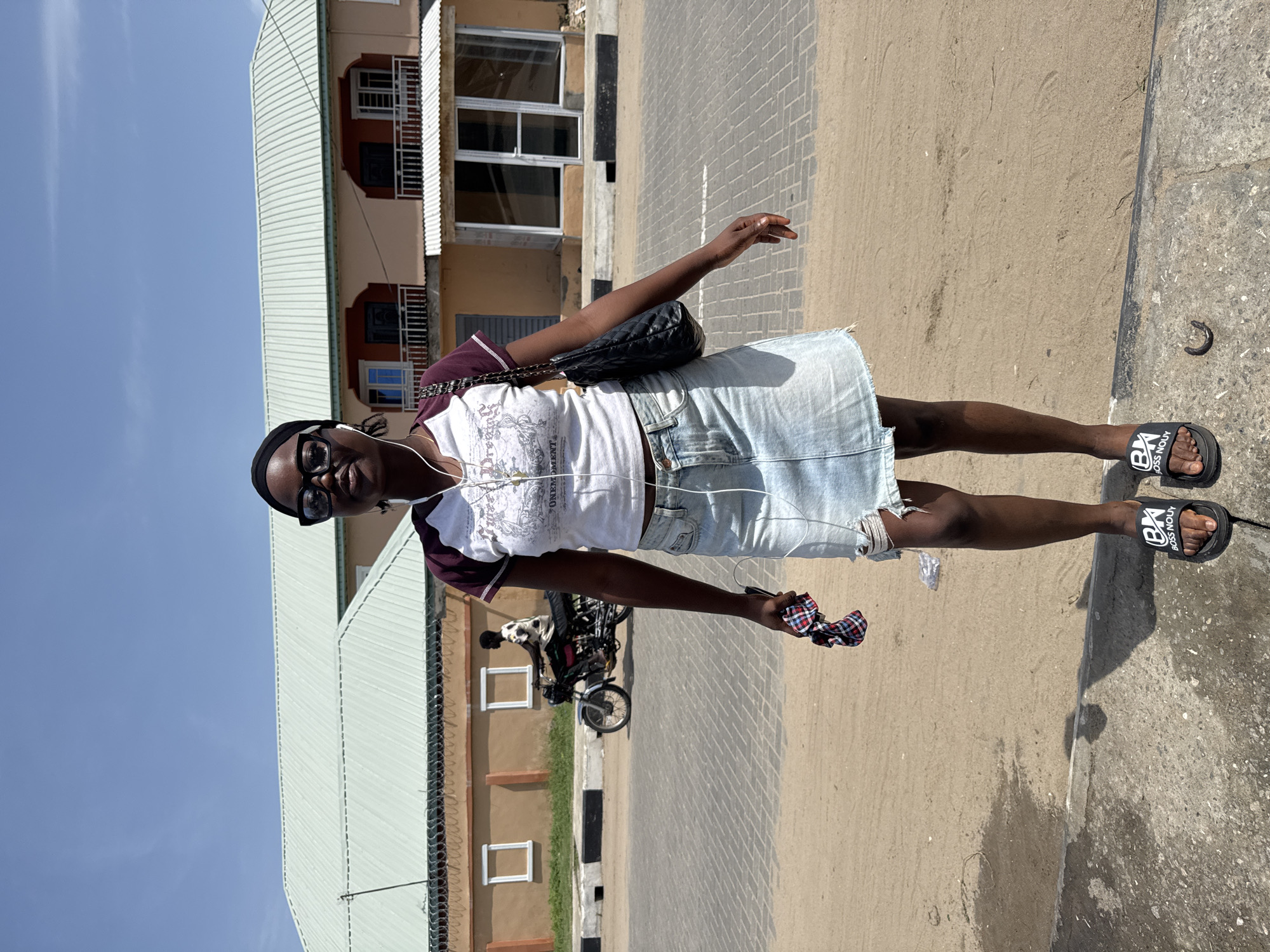 An eye-level full shot captures a slender, dark-skinned young woman outdoors in Badagry, Nigeria. She is the central figure, wearing a white t-shirt with maroon sleeves, a light blue denim skirt, black sandals with white lettering, and sunglasses. A black purse with a silver chain strap hangs from her shoulder, and white earphones are visible around her neck. She holds a scrunchie in her right hand.
Behind her is a tan building with a green roof and rectangular windows. To the left of the building is a strip of blue sky. A motorcycle is parked further behind the woman. The ground surface transitions from beige to gray cement.