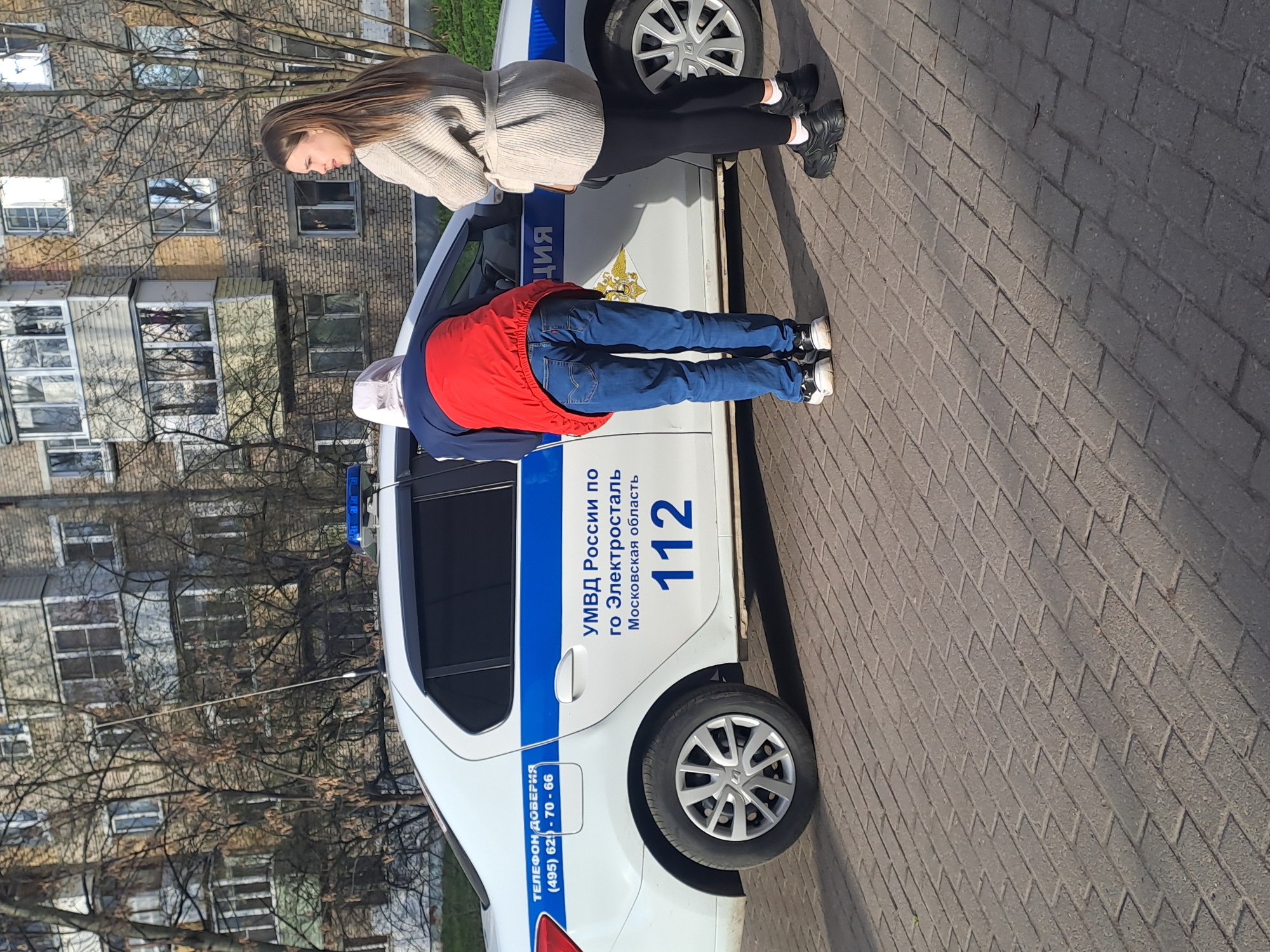 The image captures two individuals standing next to a white police car on a brick-paved surface in what appears to be an urban setting. In the background are multi-story apartment buildings and bare trees, suggesting a non-summer season.
On the left side of the police car, a woman with long brown hair is visible, looking downwards. She is dressed in a light-colored knitted sweater, black pants, and black sneakers. On the right side of the car, a second person is standing with their back to the camera, leaning against the vehicle. This individual wears a red vest over a dark blue long-sleeved top, blue jeans, and black and white sneakers, with a light-colored cap or hood on their head.
The police car is white with a prominent blue stripe along its side. Various official texts are visible on its exterior. On the rear passenger door, "УМВД России по го Электросталь Московская область" (UMVD of Russia for Elektrostal city, Moscow region) and the emergency number "112" are displayed. Further back, "ТЕЛЕФОН ДОВЕРИЯ (495) 62-70-66" (Hotline number) is written. A police emblem is also discernible. A blue flashing light is partially visible on the car's roof.
The activity appears to be an interaction or a pause next to the police vehicle. The scene is illuminated by clear daylight, casting shadows on the pavement, indicating a sunny day. The overall location cues, particularly the text on the police car, specify Elektrostal, Moscow region, Russia, as the setting.
