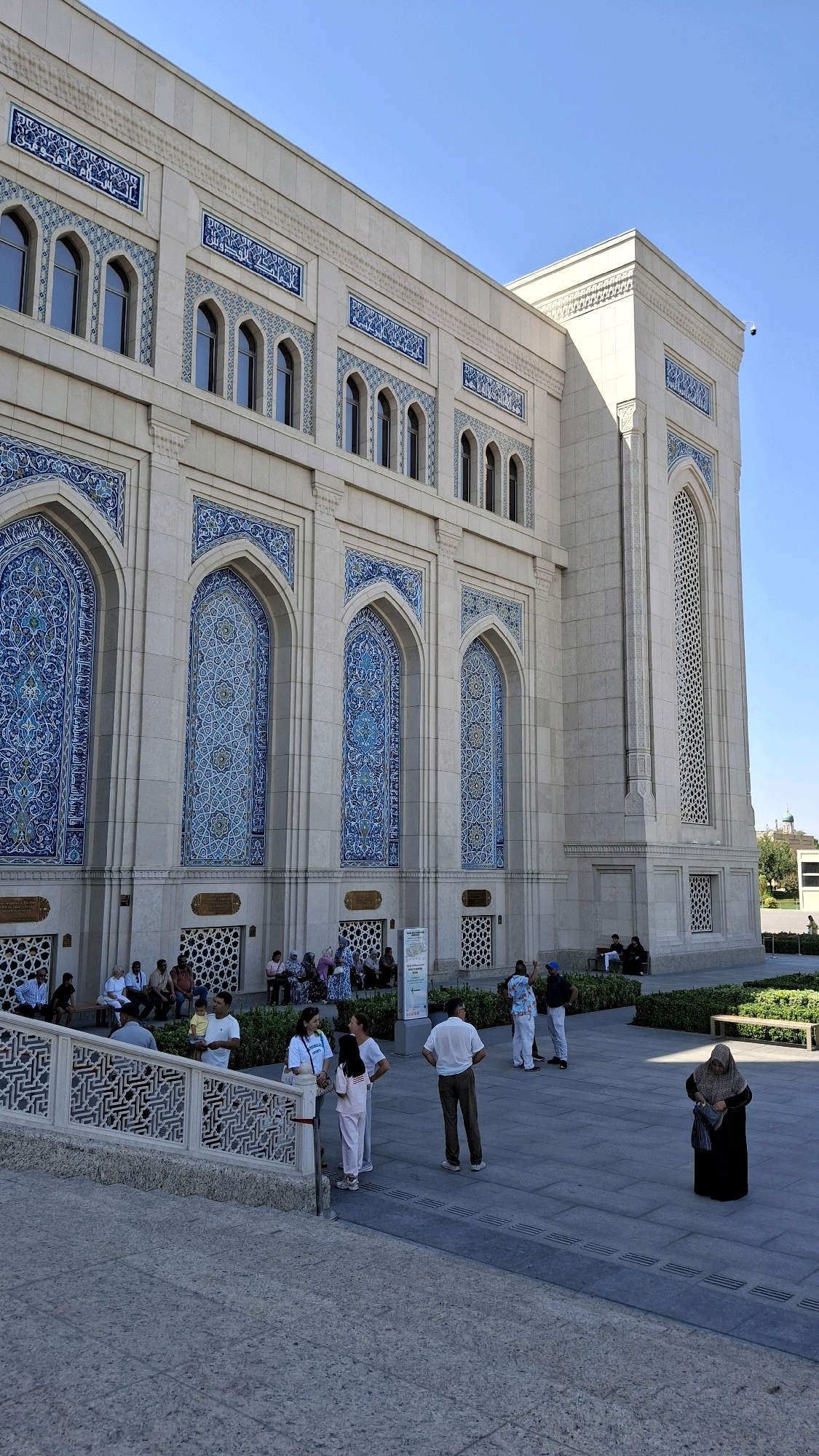 The image shows the exterior of the Minor Mosque in Tashkent, Uzbekistan, on a clear day. The mosque is a large, modern building constructed of light-colored stone, accented with intricate blue tilework in arched windows and decorative panels. A group of people are gathered outside the mosque, some sitting on benches, others standing and talking. The foreground features a textured stone pathway leading up to the mosque's entrance, with a decorative stone balustrade to the left. Several individuals, including men, women, and children, are visible in various poses, some interacting with each other, while others appear to be observing the building. One woman in the lower right corner is seen adjusting her headscarf. The overall scene depicts a public space with visitors and a clear sky overhead.
