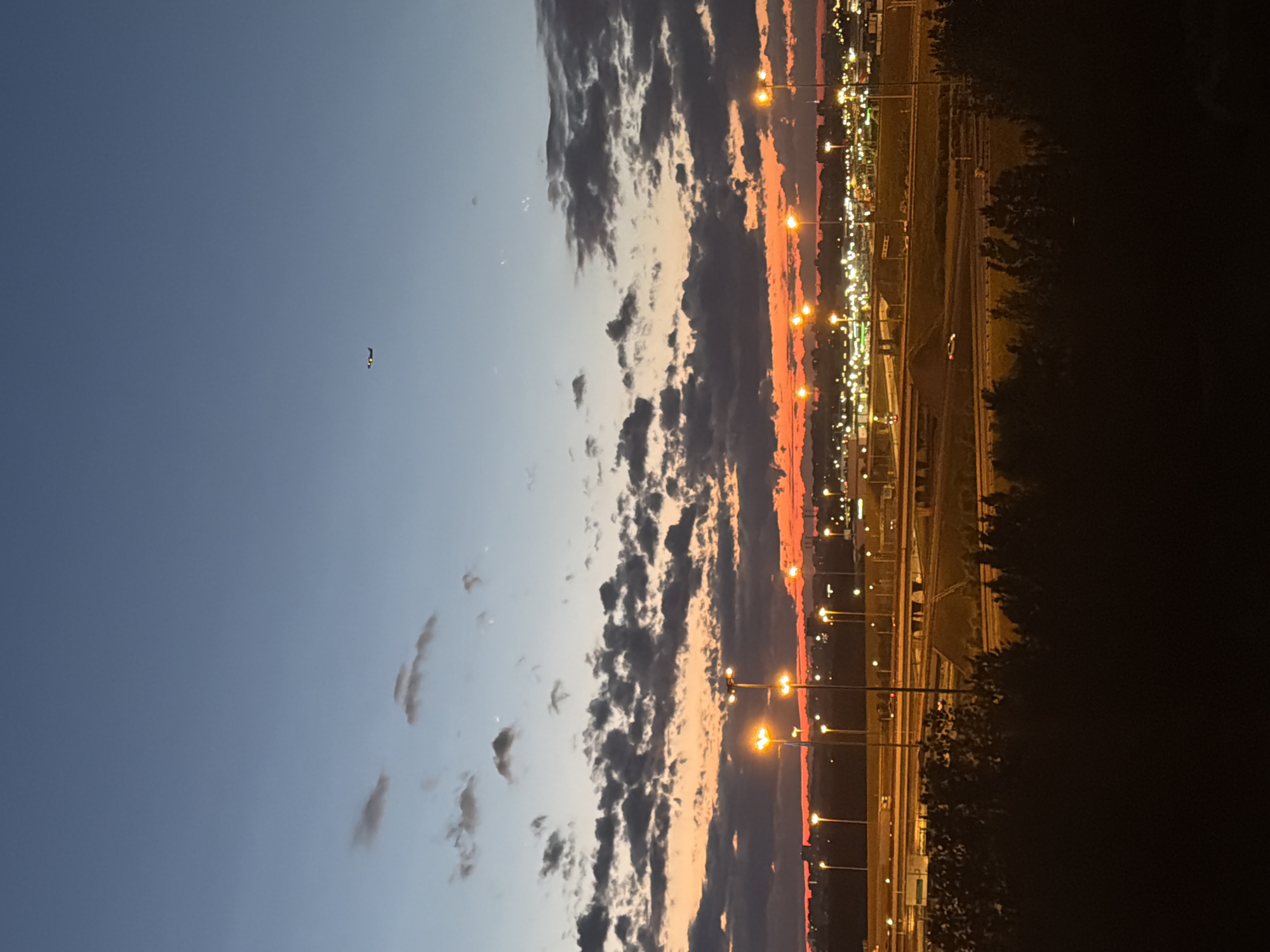 A wide shot captures a city skyline at dusk. A deep blue sky with scattered, wispy clouds transitions into a band of vibrant orange and red clouds along the horizon, indicating the setting sun. A small airplane is visible in the upper portion of the sky. Below the sky, a cityscape is illuminated by numerous streetlights and building lights, suggesting a busy urban area. Multiple elevated highways with cars are visible, crisscrossing the landscape. Dark trees in the foreground obscure the bottom of the scene.