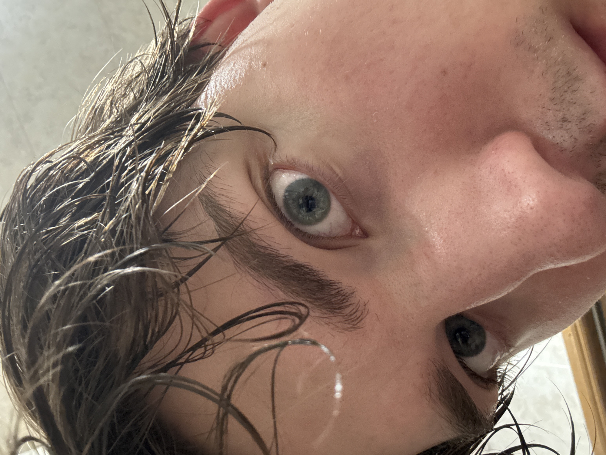 The image is a close-up, low-angle shot of a person's face, focusing on their eyes and forehead. The person has wet, dark brown hair that is falling across their forehead. Their eyes are light blue with dark pupils and are looking upwards. The skin is fair with some visible pores and light stubble on the chin and jawline. The lighting suggests an indoor setting, possibly a bathroom, with a bright light source above and to the left of the subject. There are reflections on the forehead, possibly from water droplets. No specific landmarks or identifiable features indicating the city of Dnipro, Ukraine are present in the image.