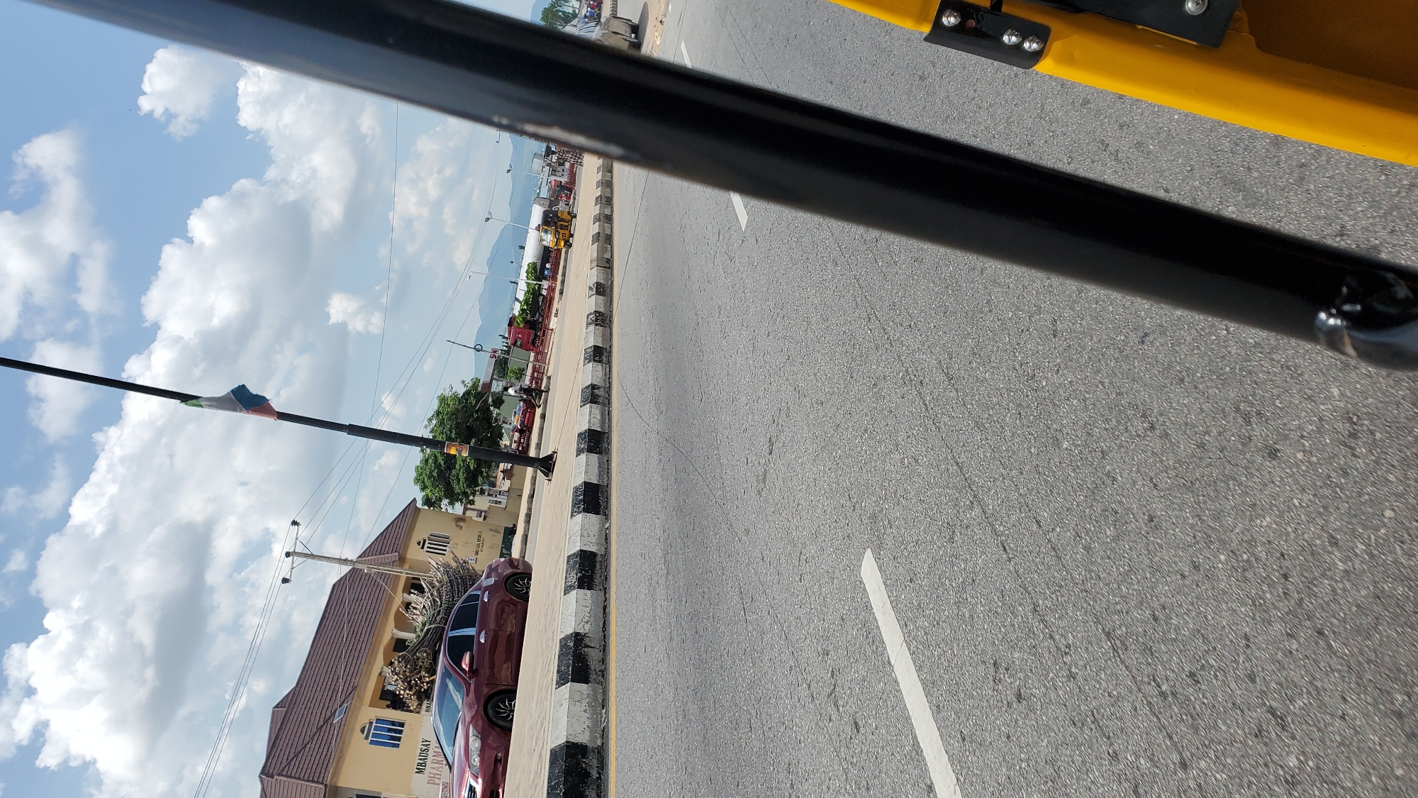 The image captures a scene on a sunny day in Jalingo, Nigeria, likely along a main road. In the foreground, a portion of a yellow vehicle, possibly a Keke Napep, is visible on the right, with a black pole from the vehicle structure running diagonally across the frame. The road surface is asphalt with a white lane marking.
To the left of the road, a maroon sedan is parked. Behind the sedan, a multi-story building with a brown roof and light-colored walls stands. Text on the building, partially visible, reads "MBAUSAY PHARMA". A cluster of tall, bundled stalks, likely sugarcane or similar produce, is loaded onto the roof of the sedan.
Further down the road, a flag with green, white, and blue bands is attached to a black pole. Beyond this, there are other vehicles, including a red truck and a yellow tricycle-like vehicle, and what appears to be a gas station with visible pumps. In the distance, hills and mountains are visible under a bright sky with scattered white clouds. A few individuals are present on the sidewalk and near the vehicles. The overall impression is a typical urban roadside scene in Nigeria, with commercial activity and transportation.